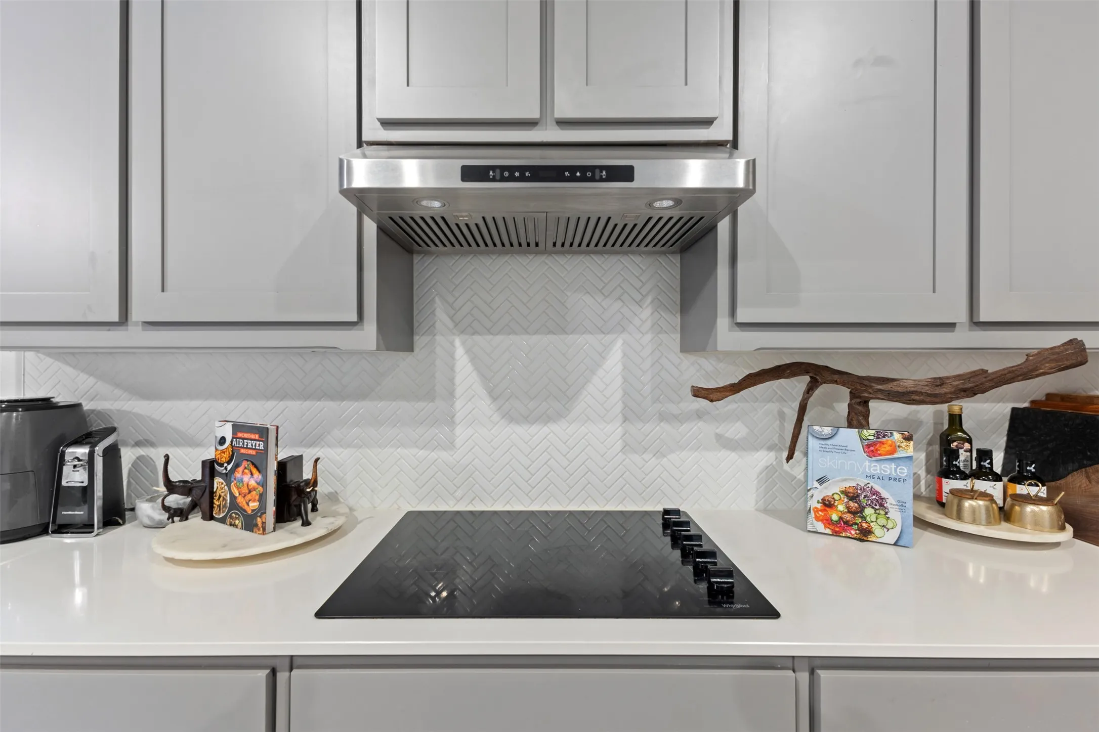 Sleek cooktop with a stainless steel range hood, light gray cabinetry, and a white herringbone tile backsplash, set against a solid surface countertop