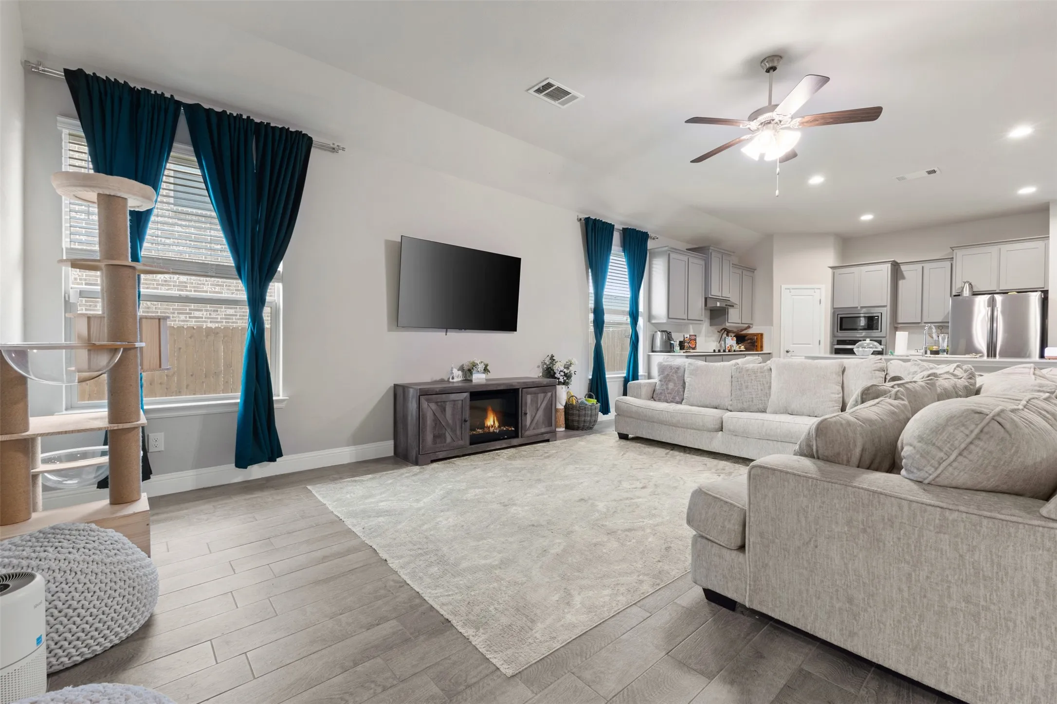 Open-concept living area featuring light wood-finish flooring, a gas fireplace with dark cabinetry surround, wall-mounted flat-screen television, ceiling fan with integrated lighting, recessed ceiling lights, and expansive windows dressed with deep blue drapery