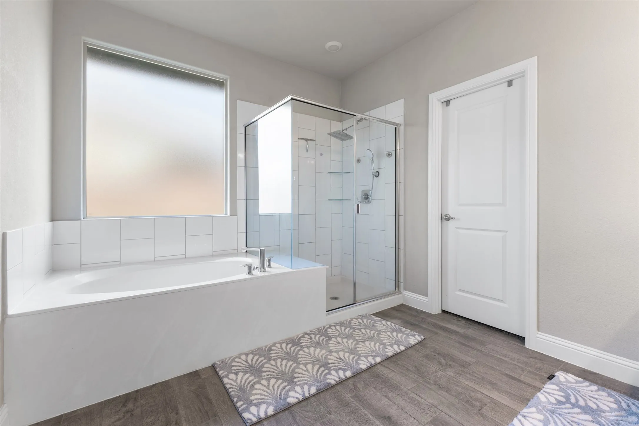 Built-in bathtub with white tile surround, adjacent glass-enclosed shower with white subway tile, and wood-finish flooring