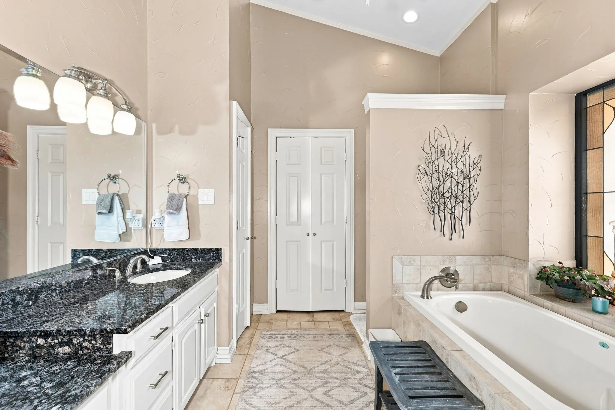 Double vanity with dark stone countertop, built-in bathtub with tiled surround, and vaulted ceiling