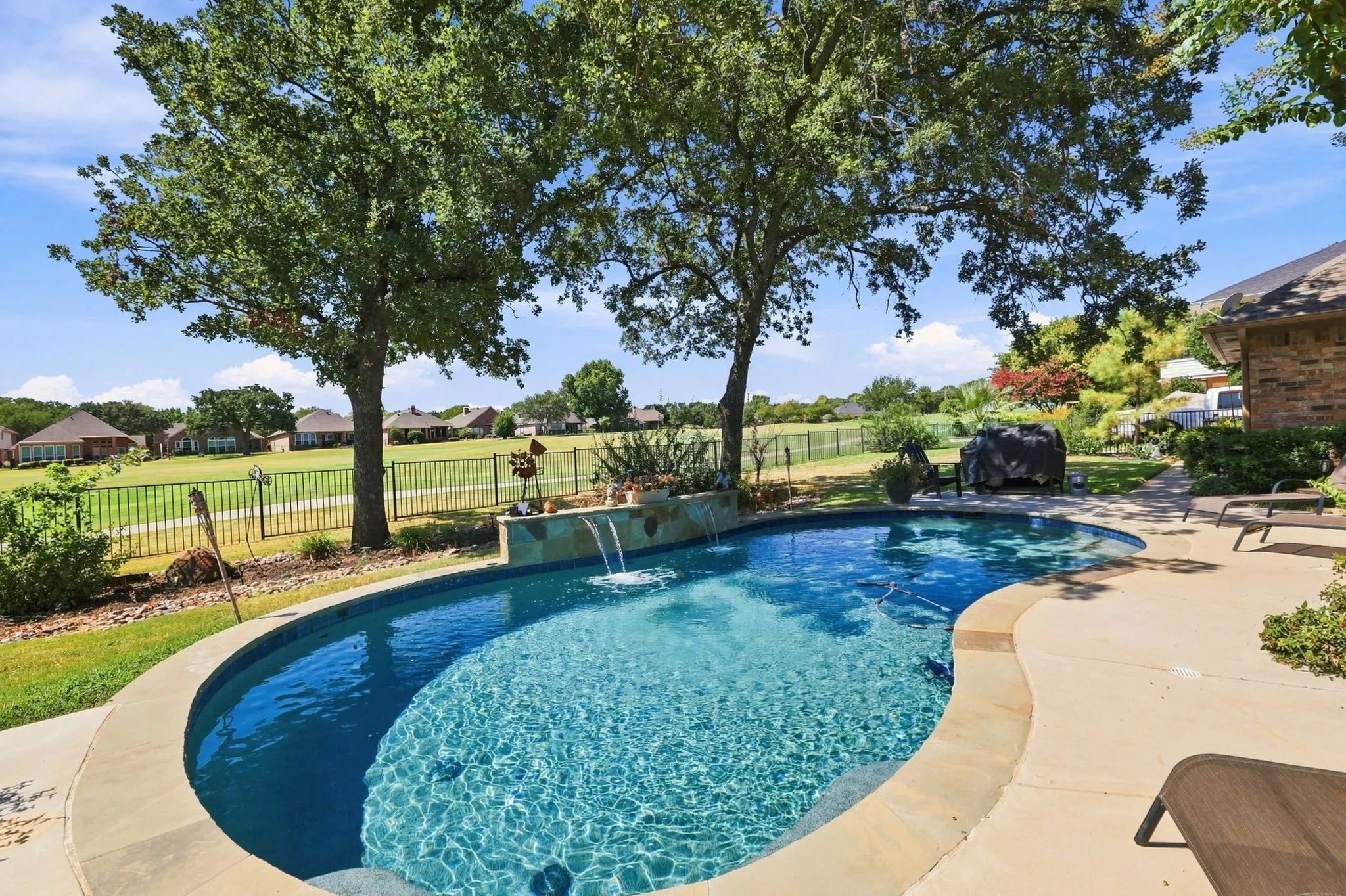 Freeform swimming pool with stone coping, integrated water features, expansive patio, and mature trees