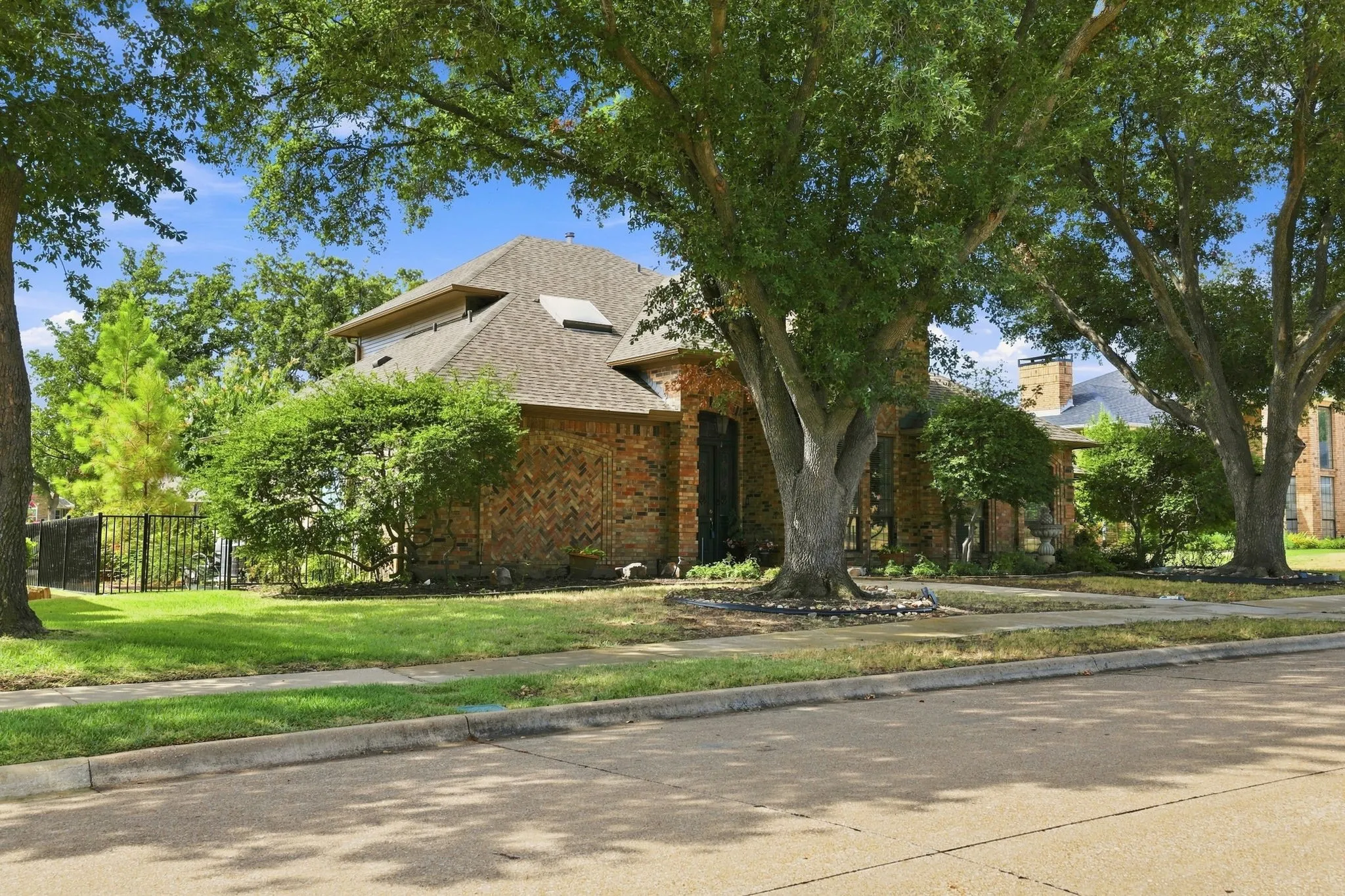 Brick exterior with a gabled shingle roof, mature trees, and a decorative wrought iron fence