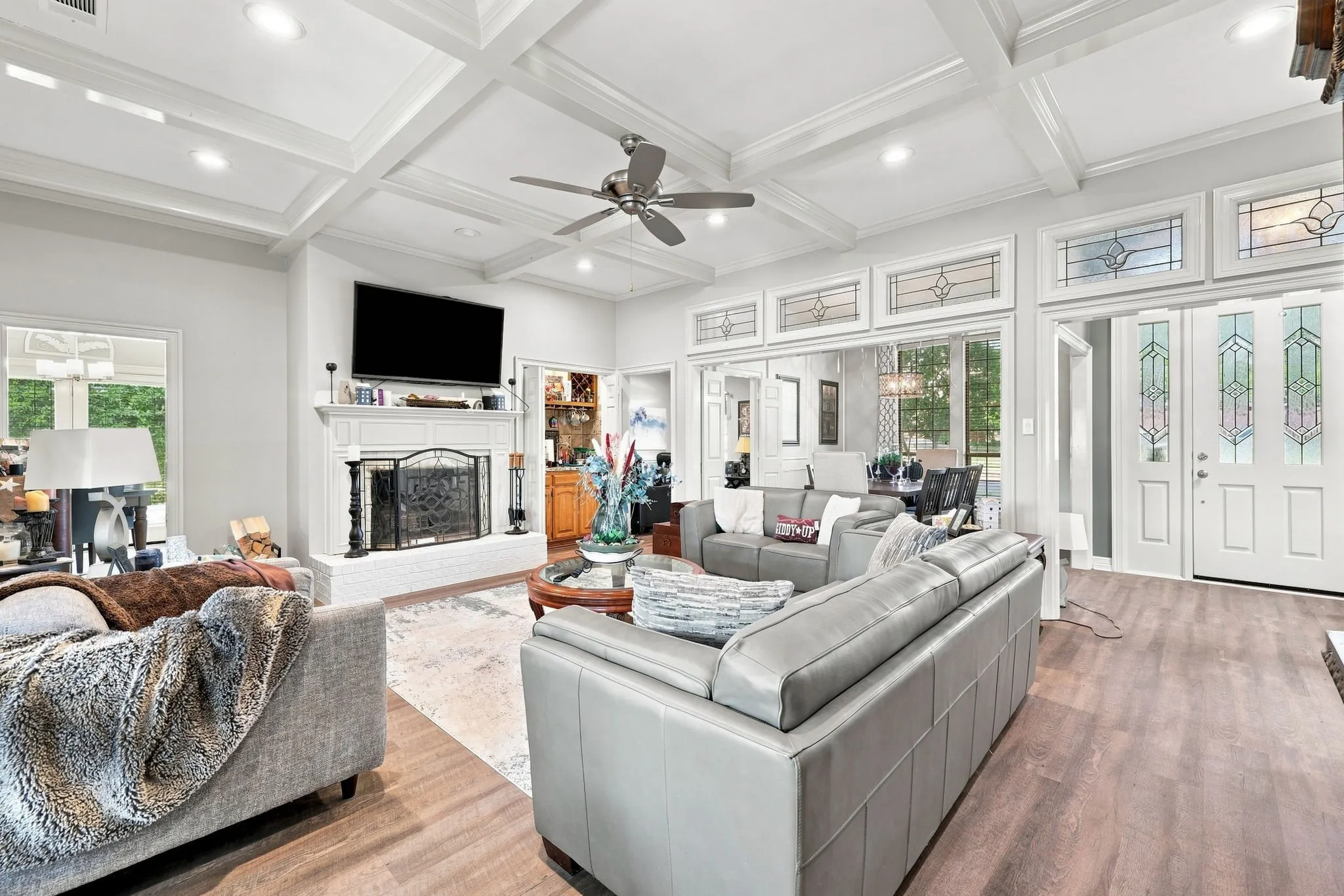 Coffered ceiling with recessed lighting, wood-finish flooring, white brick fireplace with mantel, double entry doors with decorative glass panels, and transom windows with decorative glass