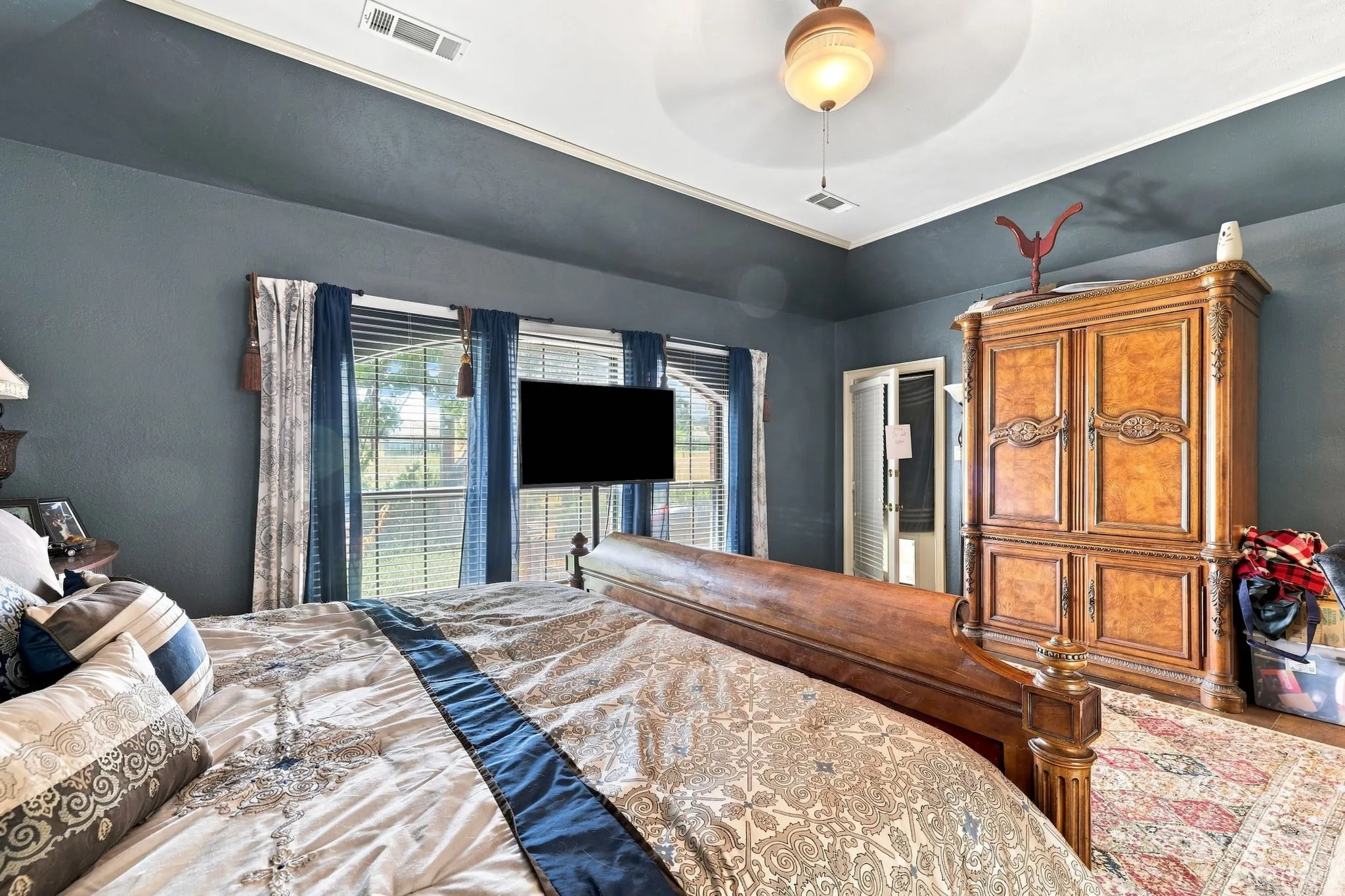 Spacious primary bedroom featuring slate blue walls, dual glass doors with natural light, wall-mounted flat-screen television, wood-finish cabinetry with decorative hardware, and a ceiling-mounted light fixture