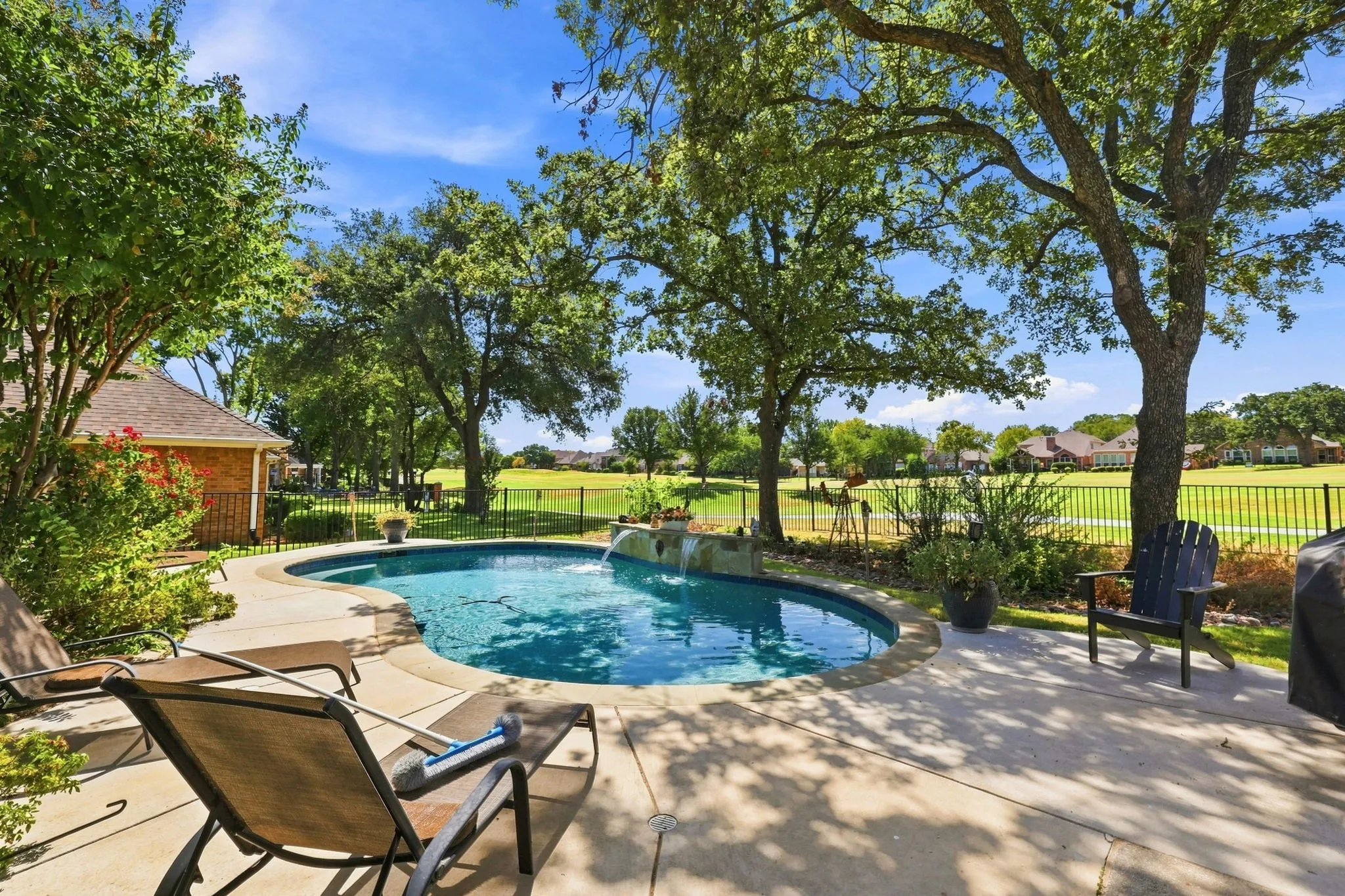 Curved swimming pool with water features, surrounded by a concrete patio, mature trees, and a black metal fence bordering a green landscape