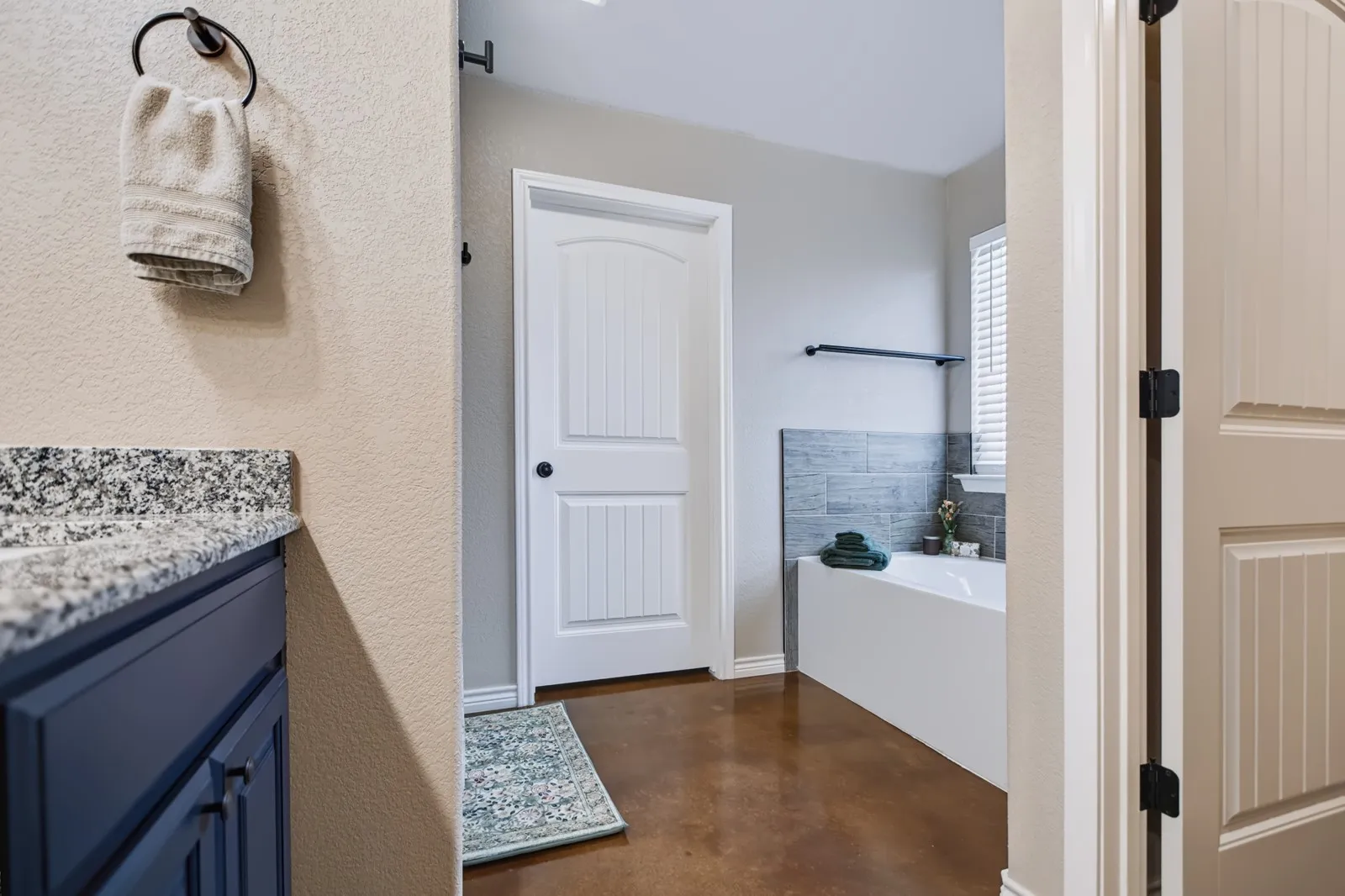 Granite-style countertop, cabinetry with dark finish, wood-finish flooring, built-in tub, and tiled wall accent