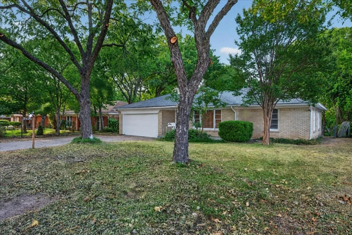 Light brick exterior, attached garage, mature trees, manicured shrubbery, and a sloped roofline
