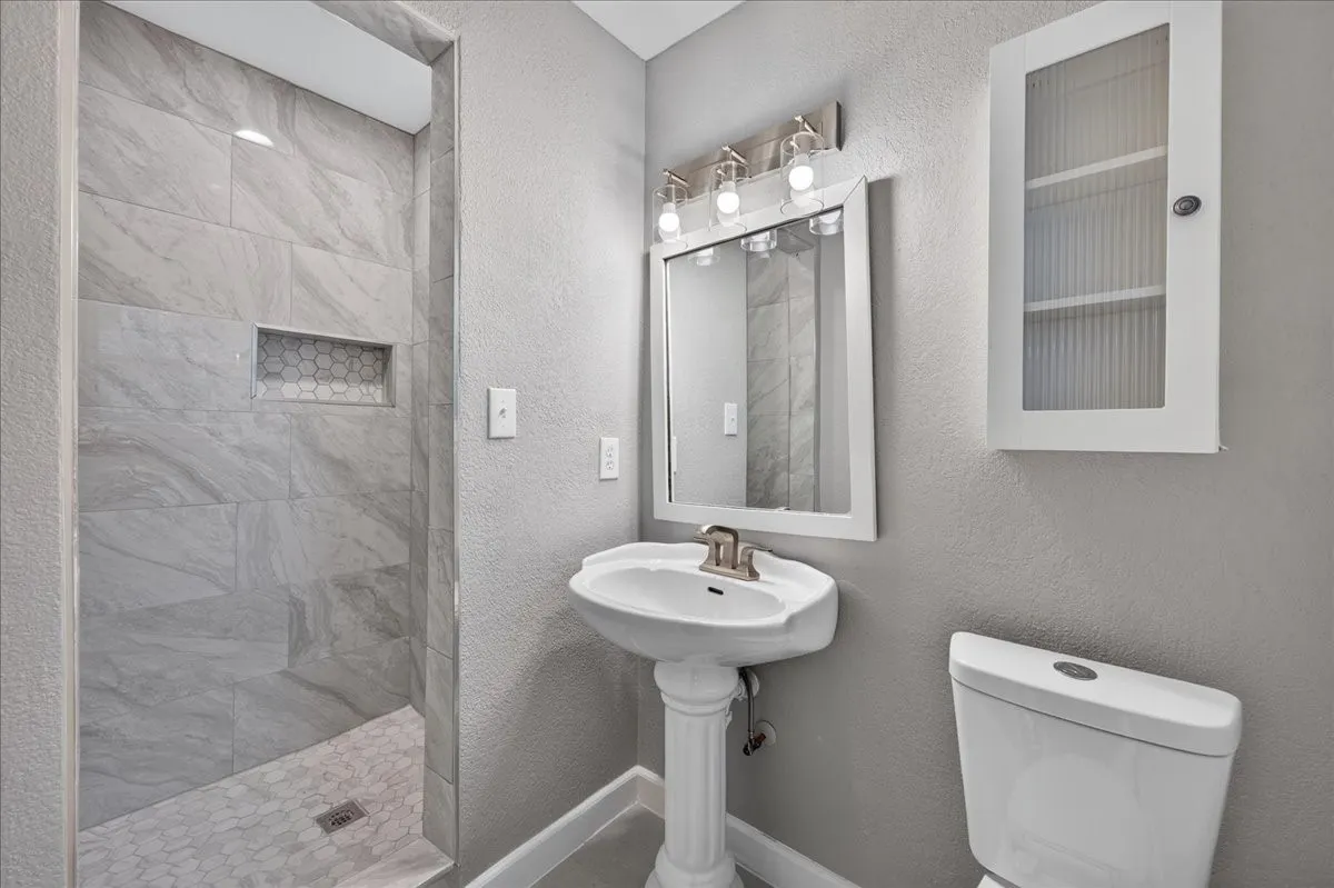 Walk-in shower with large format tile and hexagon tile niche, pedestal sink, wall-mounted mirror with vanity lighting, and wall-mounted storage cabinet
