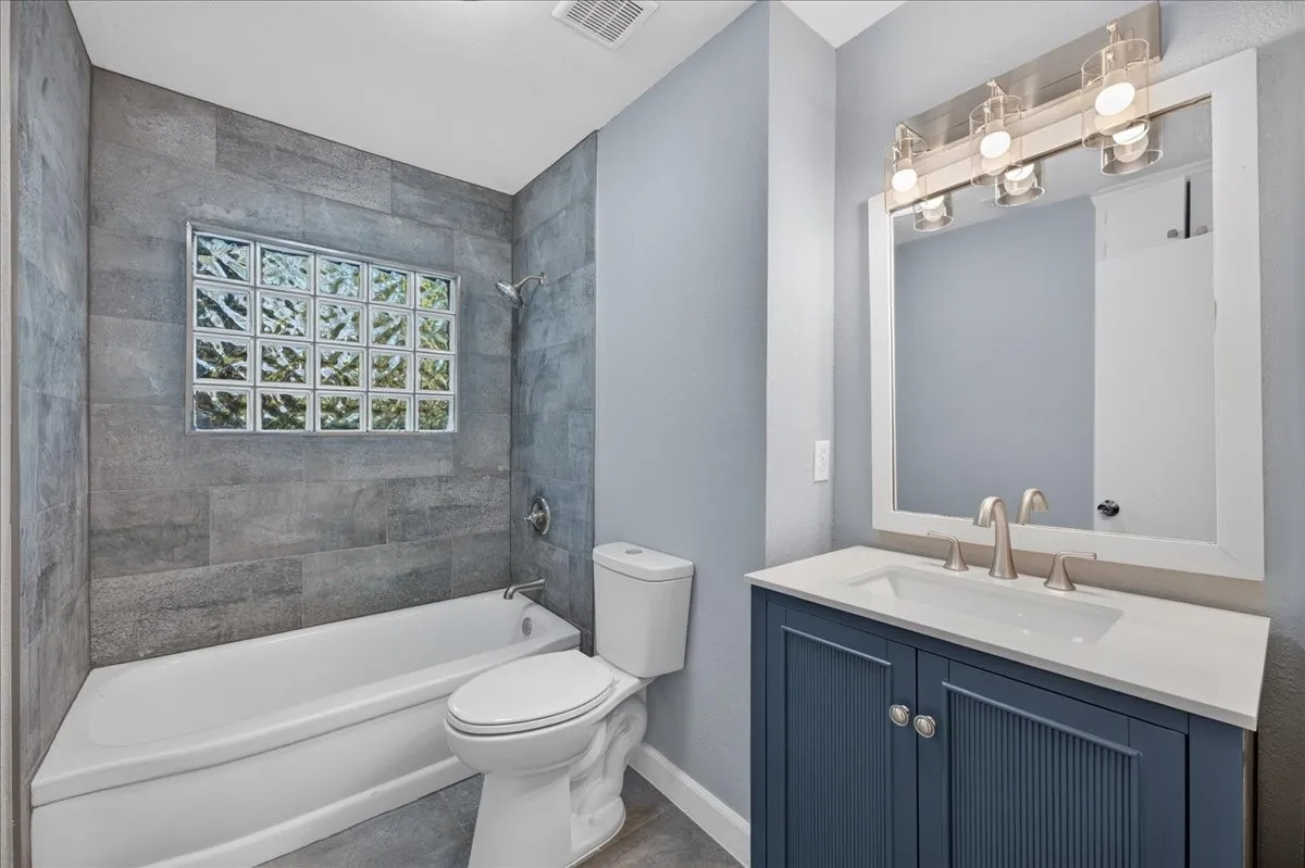 Bathroom featuring a vanity with a white countertop and blue cabinetry, a wall-mounted mirror, brushed nickel faucet, and a tiled shower with a glass block window