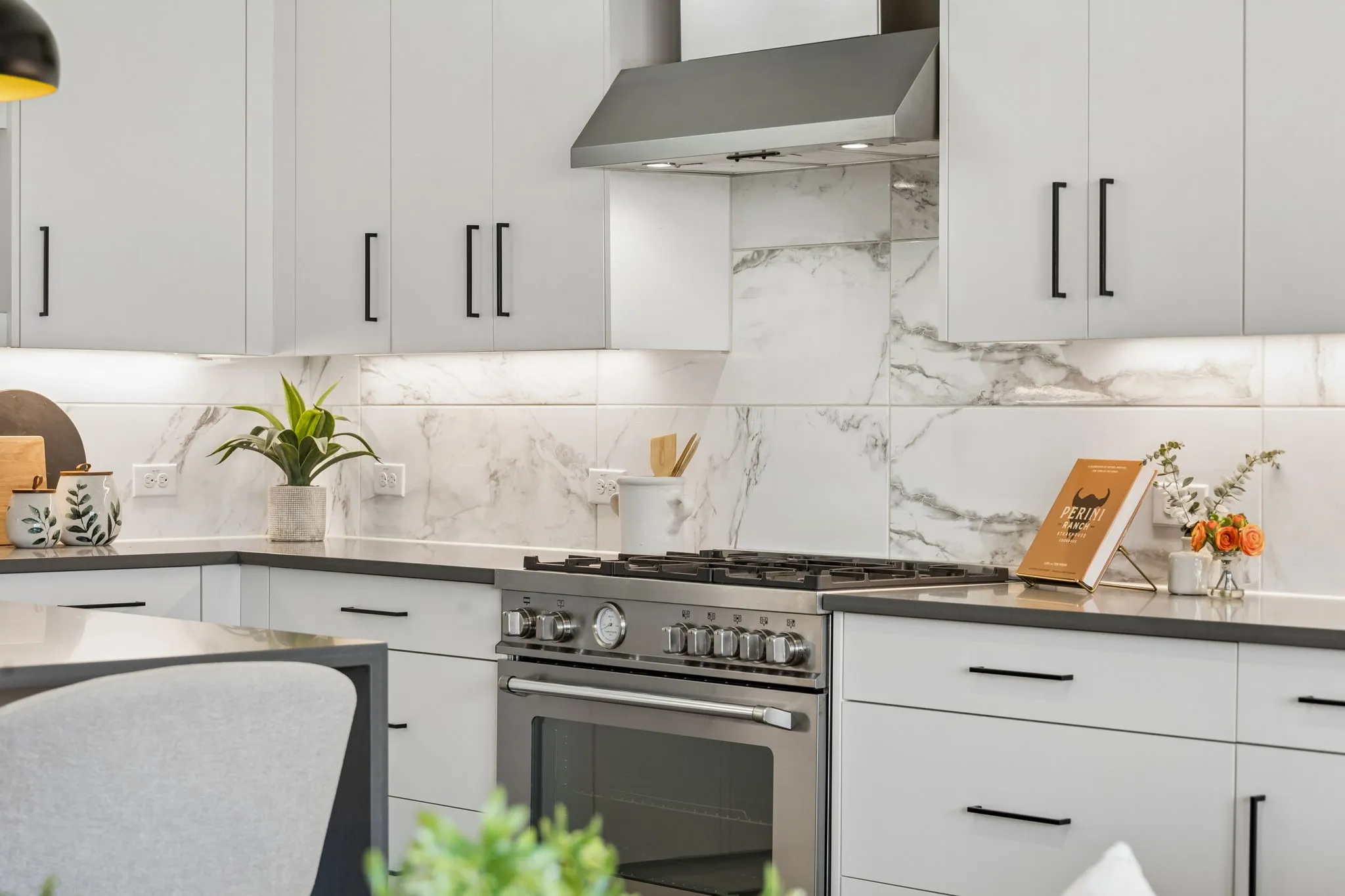 White shaker-style cabinetry with matte black hardware, stainless steel range with hood, stone countertops, and a white marble-patterned tile backsplash