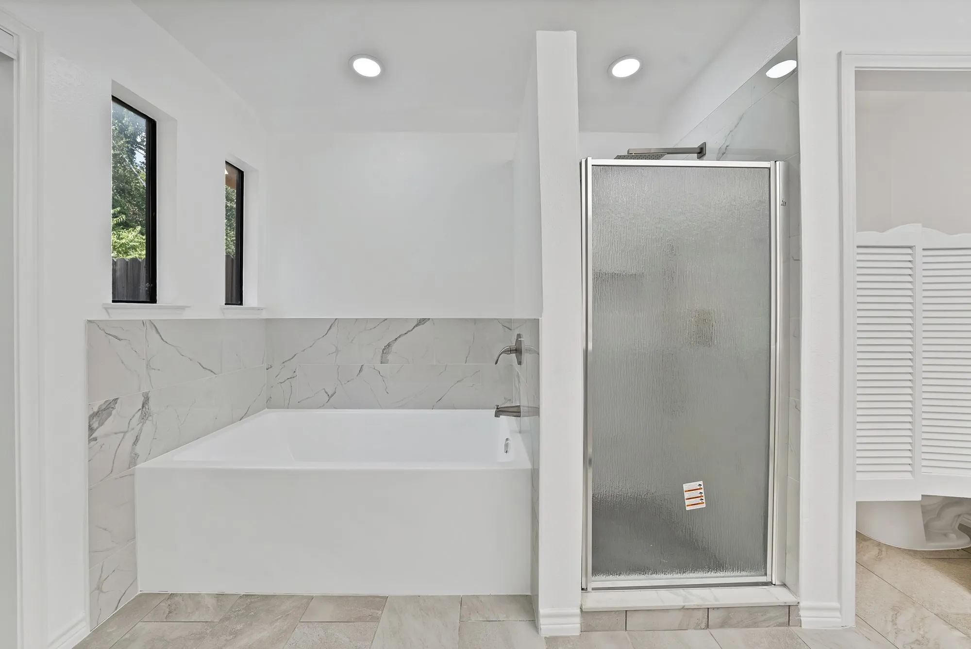 Built-in bathtub with a tile surround, glass-enclosed shower, two vertical windows, and recessed lighting