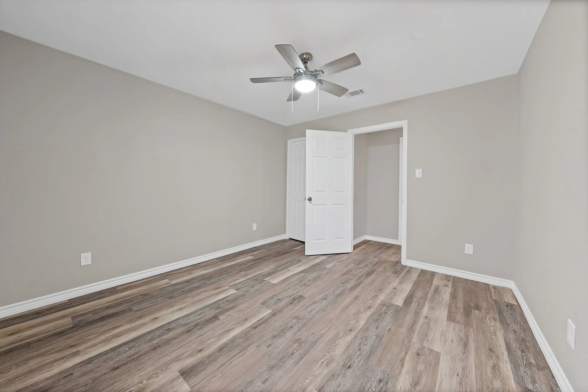 Spacious room featuring wood-finish flooring, light gray wall paint, a ceiling fan with integrated lighting, and white trim