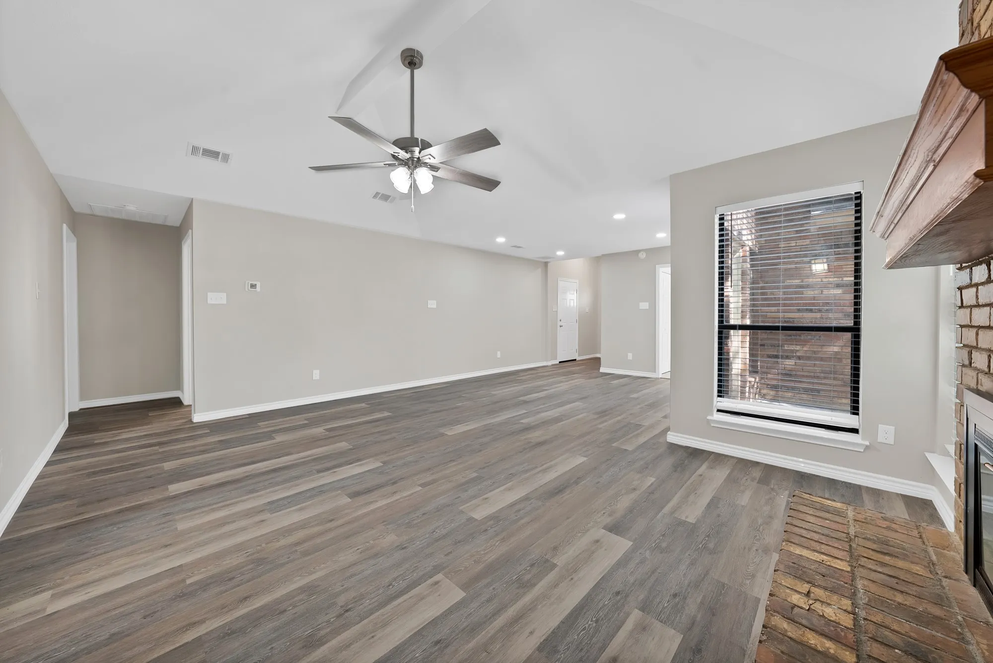 Spacious living area featuring wood-finish flooring, vaulted ceilings, a ceiling fan with integrated lighting, a brick fireplace, and a window with blinds