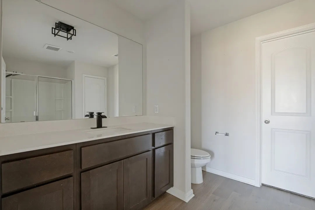 Spa-inspired primary bathroom showcasing a dark wood dual-vanity with ample storage, white quartz countertop, vessel sink with matte black faucet, large framed mirror with integrated lighting, tile flooring, and separate water closet with white six-panel door