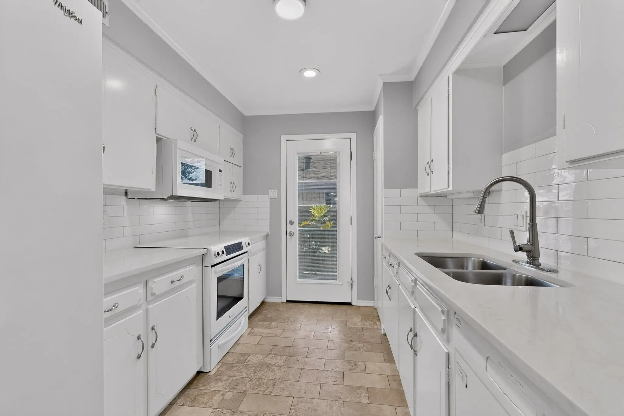 White cabinetry, light-toned countertops, subway tile backsplash, stainless steel double basin sink, and a glass-paneled exterior door