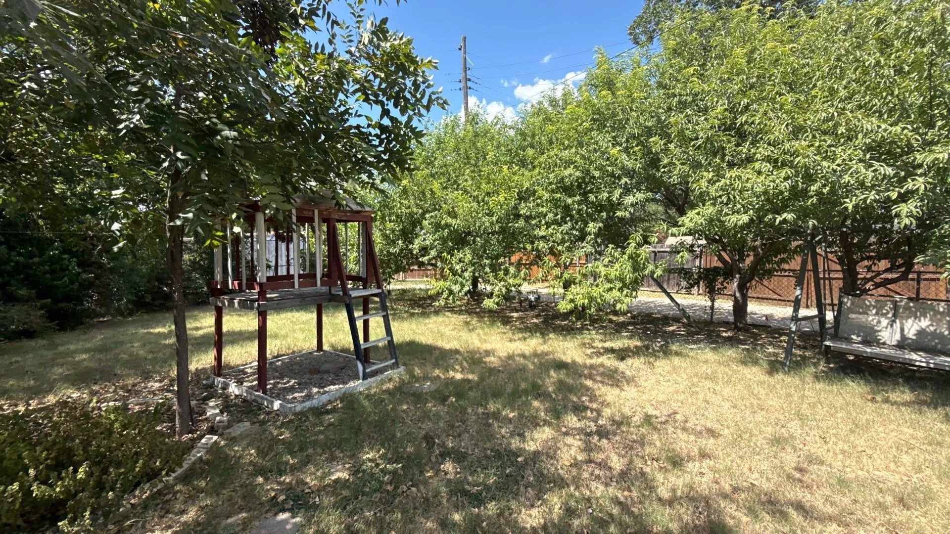 Expansive green space featuring mature trees, a multi-level wooden play structure, and a wooden swing
