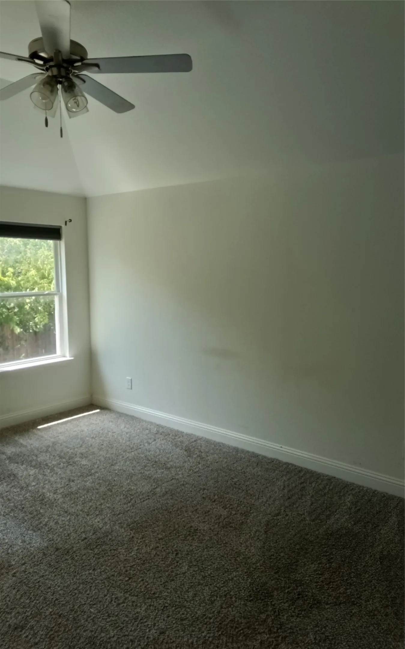 Vaulted ceiling with a ceiling fan, window with a dark roller shade, neutral wall paint, and gray carpeting