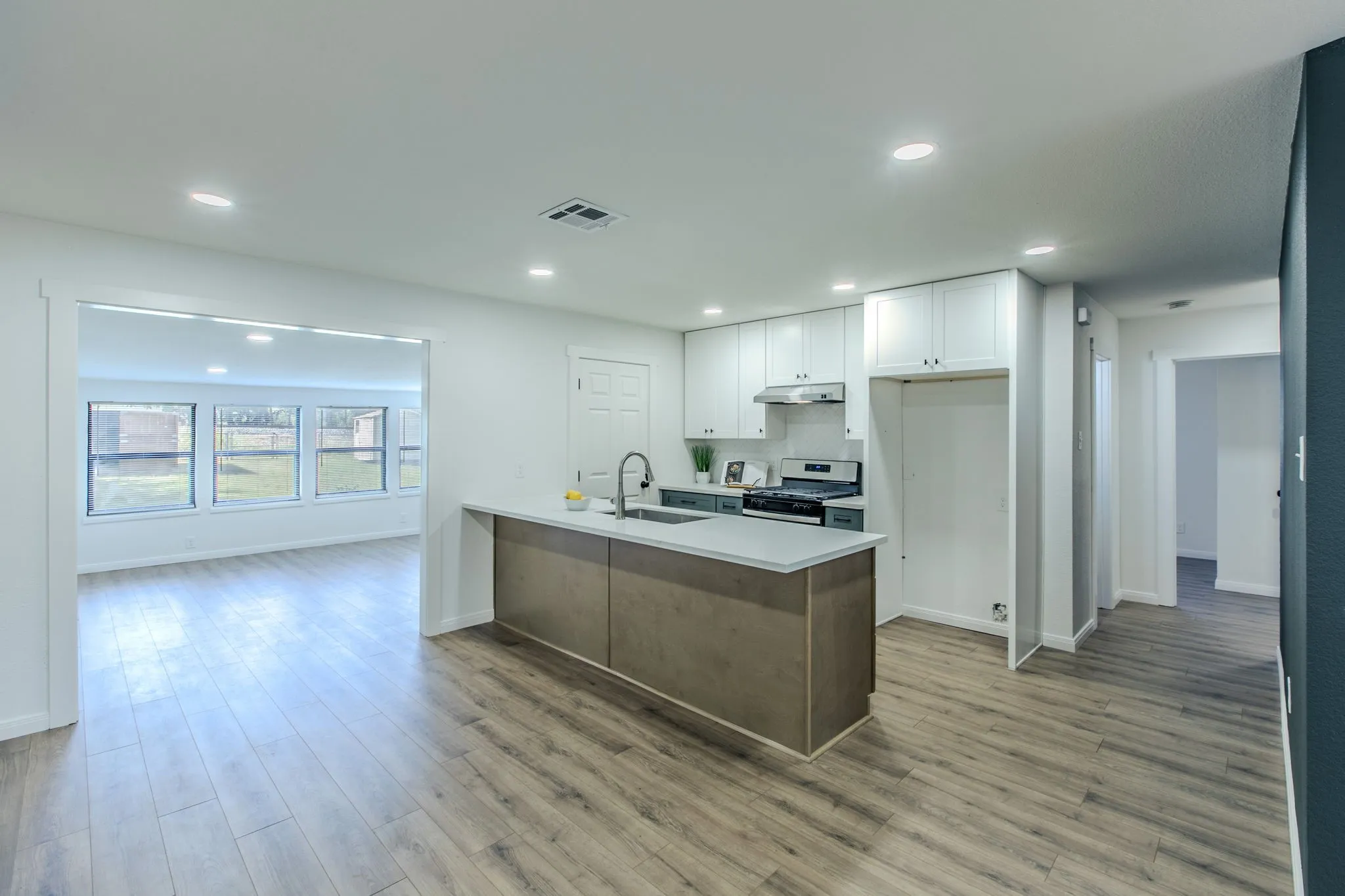 Modern open-concept kitchen and living area featuring a wood-finish plank floor, stainless steel appliances, white cabinetry, a large island with wood-finish base and countertop seating, recessed lighting, and abundant natural light from multi-pane windows