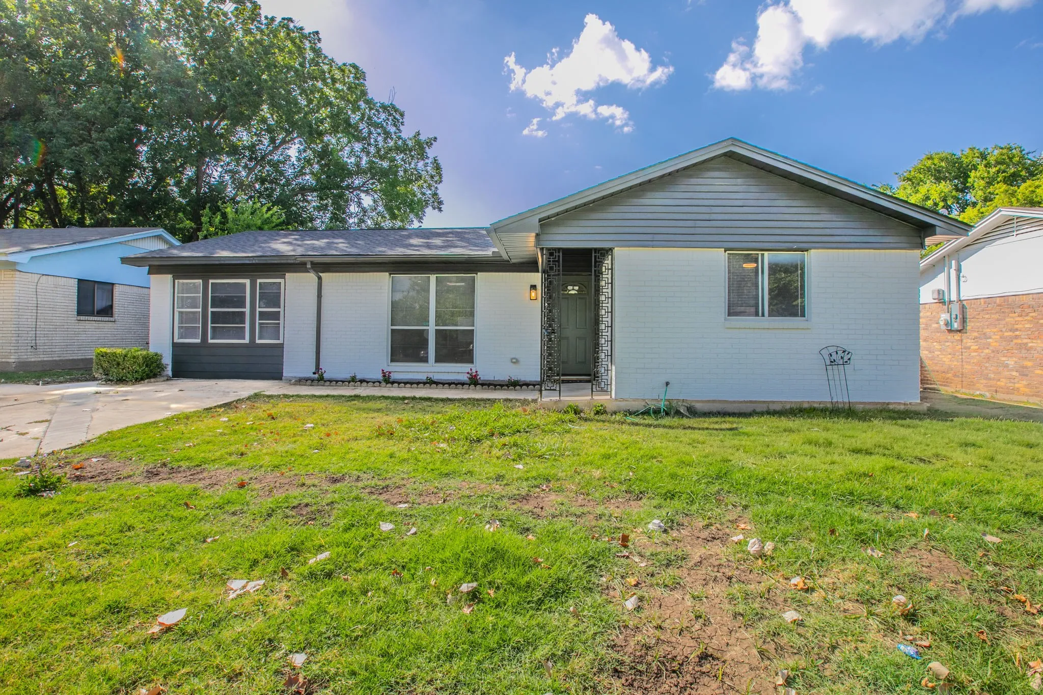 Modern ranch home featuring a refreshed exterior with light siding and contemporary dark-trimmed windows, mature landscaping with established trees providing natural shade, a spacious front lawn with level grading, and clean architectural lines with an updated front entry design