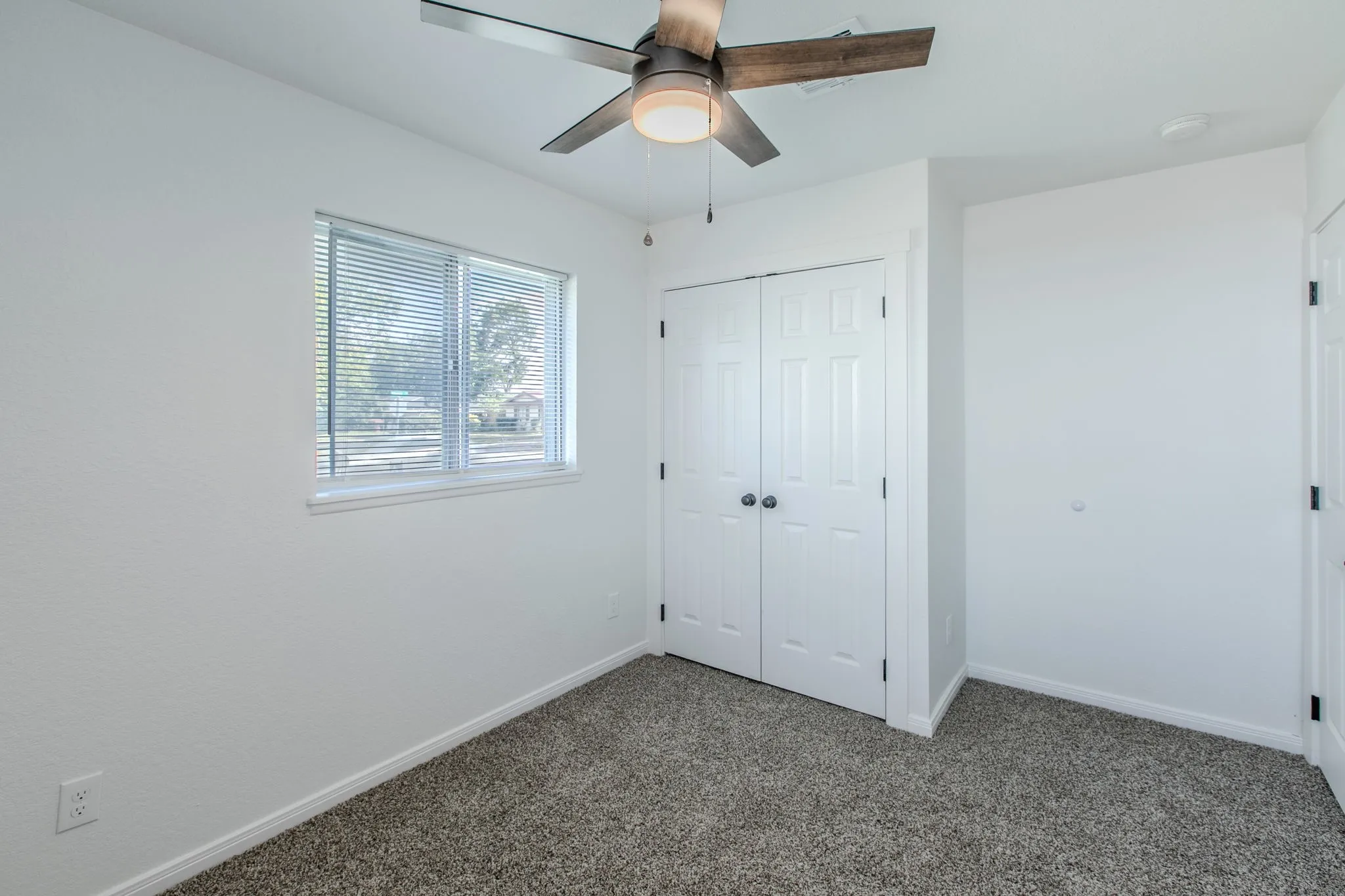 Textured carpeting, a ceiling fan with light fixture, double panel closet doors, and a window with blinds