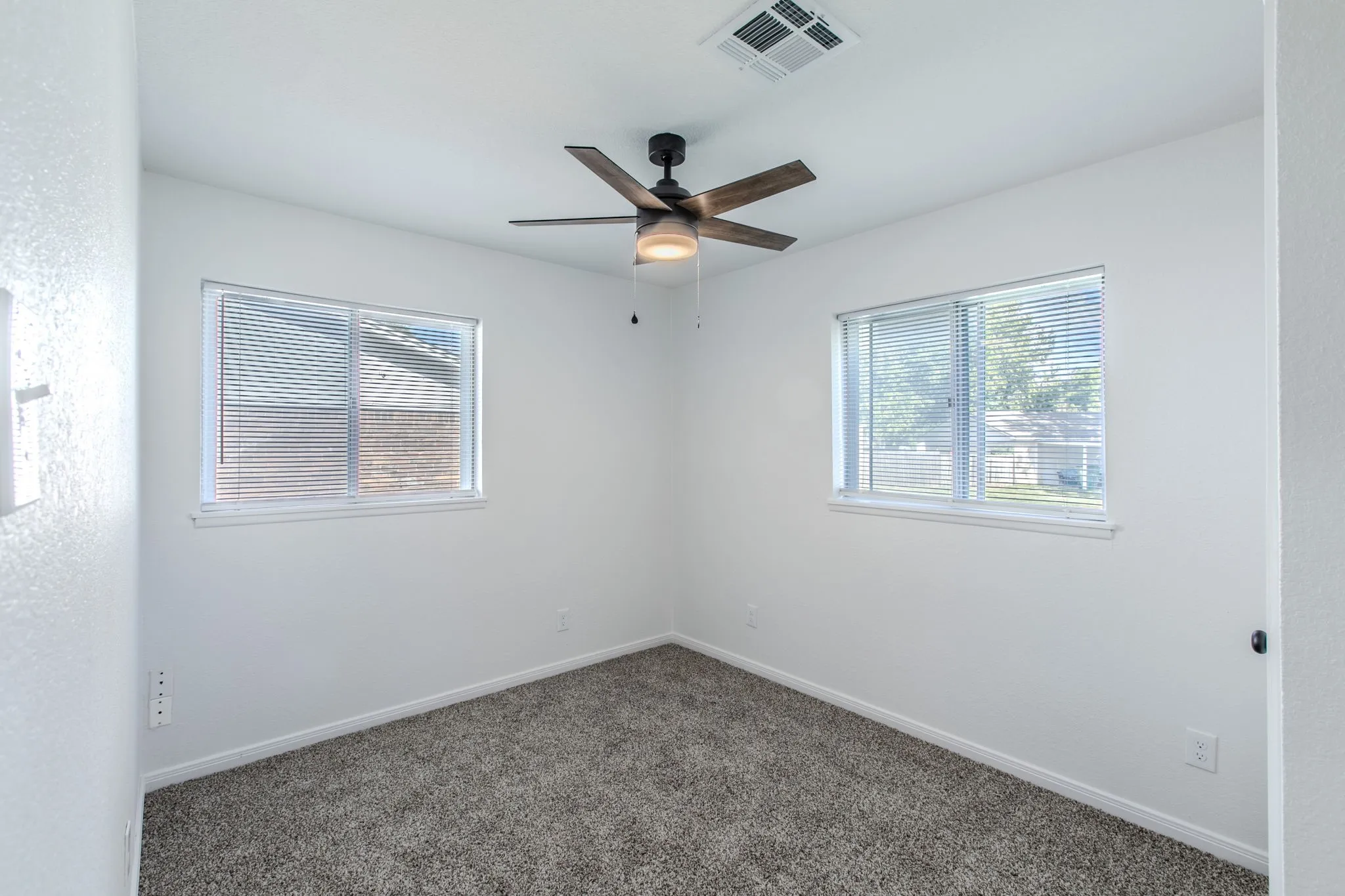 Two windows with blinds, wood-finish ceiling fan with integrated lighting, textured white walls, and gray carpet
