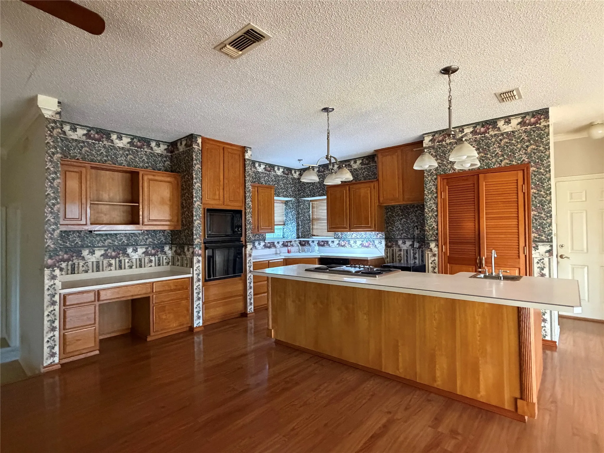 Kitchen featuring wood-finish flooring, built-in double ovens, extensive wood cabinetry, and a large island with a sink and pendant lighting