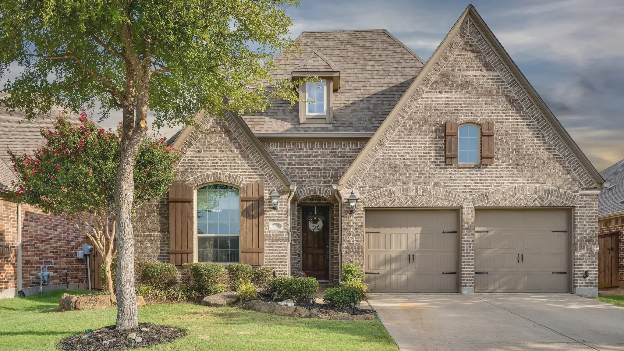 Brick exterior with multiple gables, arched entry, two-car garage, dormer window, and mature landscaping