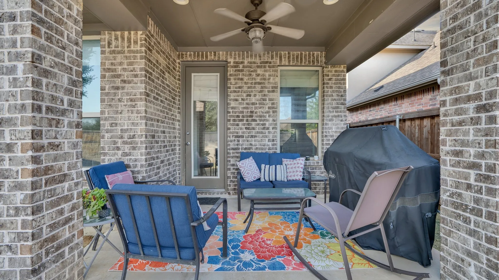 Covered porch with brick exterior, ceiling fan, and built-in seating area featuring blue Adirondack-style chairs and metal side tables