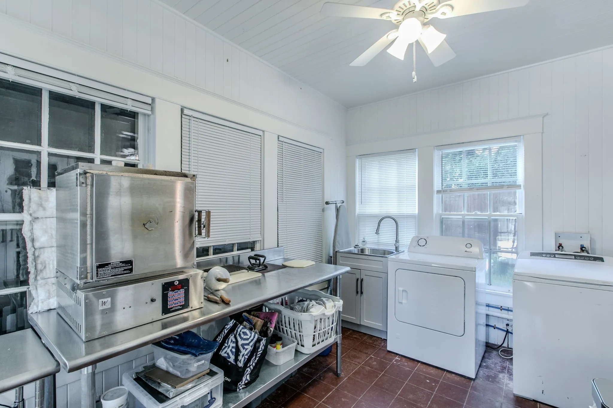 Spacious laundry room featuring stainless steel front-load washer and dryer, ample countertop workspace with storage below, white cabinetry, terra cotta tile flooring, and abundant natural light from multiple windows
