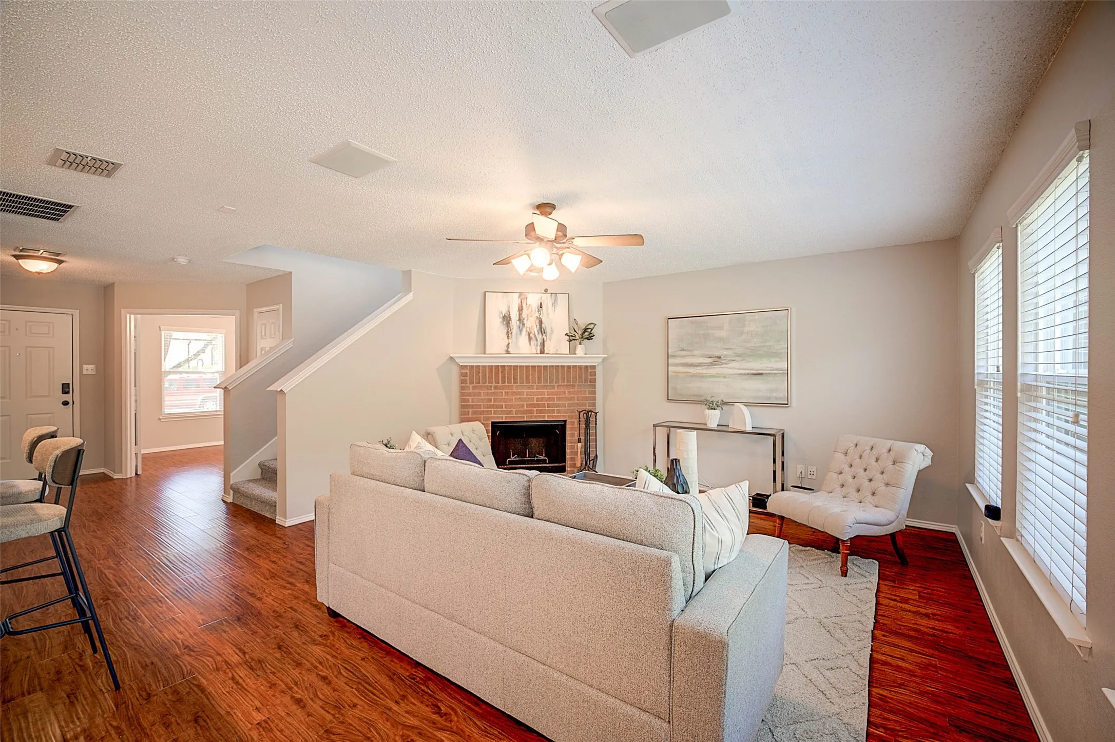 Wood-finish flooring throughout, brick-surround fireplace with a white mantel, ceiling fan with integrated lighting, and multiple windows with blinds