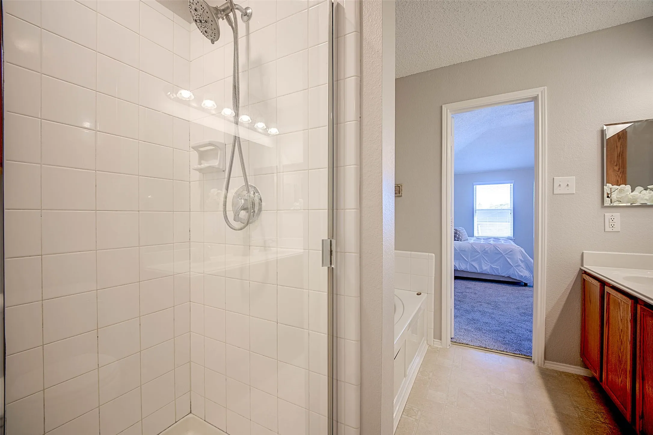 Tiled shower with glass enclosure and chrome fixtures, built-in soaking tub, wood-finish vanity with countertop, and framed mirror