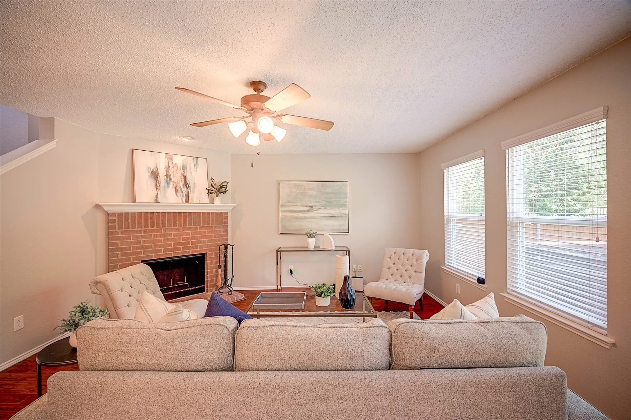 Living area featuring a brick fireplace with a white mantel, ceiling fan with integrated lighting, wood-finish flooring, and multiple windows with blinds