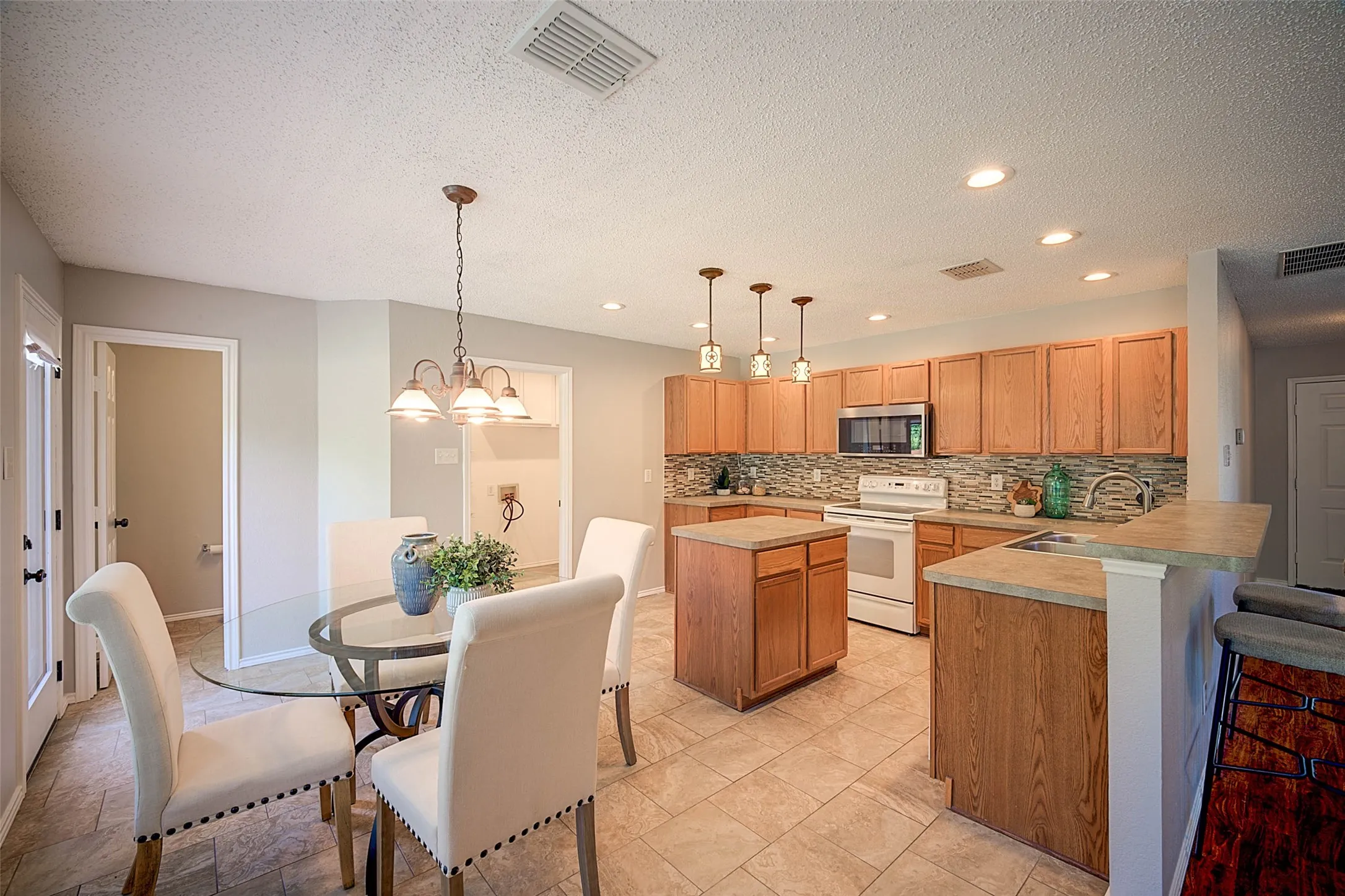 Open-concept kitchen and dining area featuring warm wood cabinetry, tile countertops, stainless steel appliances, recessed ceiling lighting with pendant fixtures, and tile flooring throughout the space