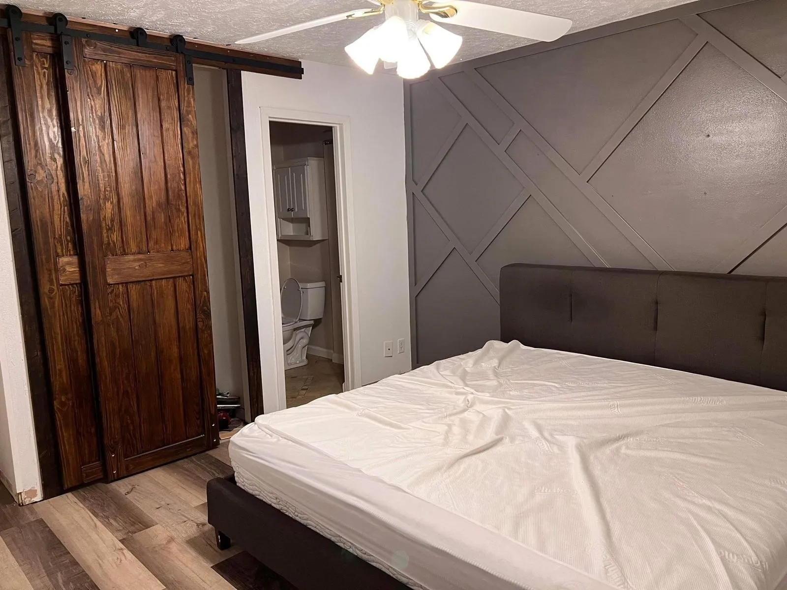 Wood-finish flooring, accent wall with geometric paneling, ceiling fan with light fixture, and wood barn doors