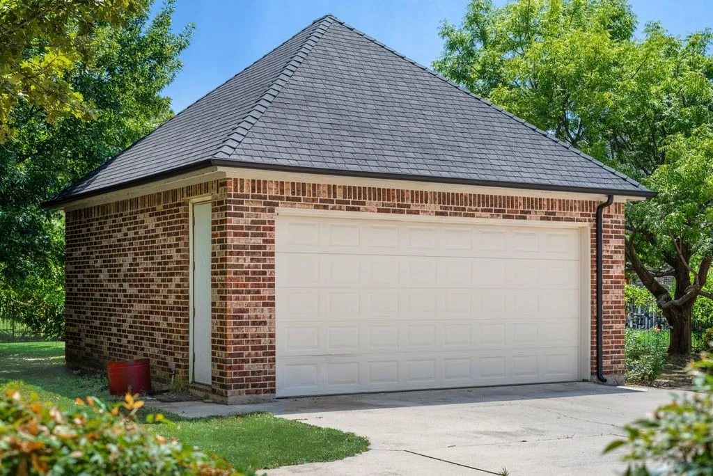 Brick-clad garage structure featuring a hip roof with dark shingles, a white panel garage door, a side entry door, and dark gutters