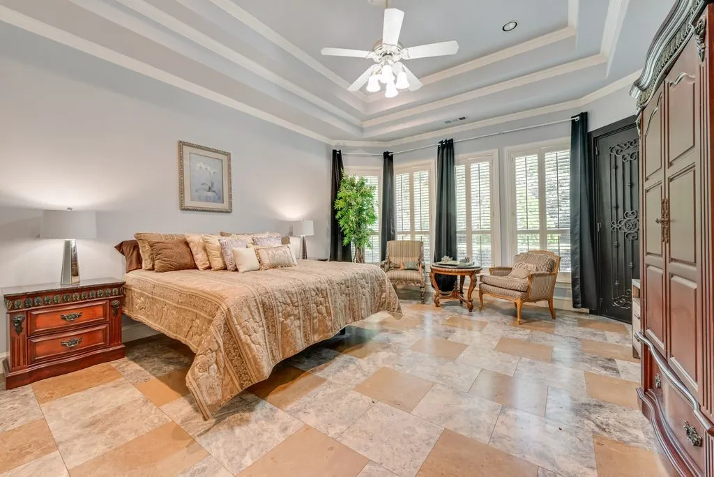 Spacious Primary bedroom featuring tray ceilings, light-tone tile flooring, and multiple windows with plantation shutters.