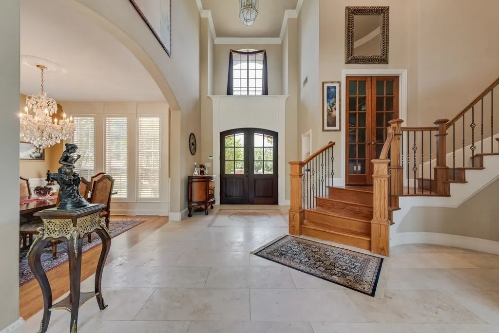 Two-story foyer featuring a grand wooden staircase with wrought iron balusters, light-toned tile flooring with an ornate inlay, and double arched entry doors.