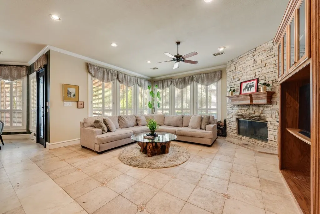 Living area featuring light-toned tile flooring with decorative inserts, a stone-front fireplace with a wood mantle, and an extensive bank of windows with plantation shutters.