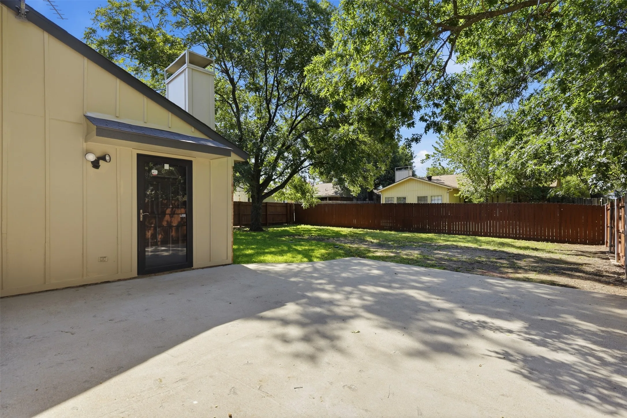 Concrete patio with a side entrance featuring a screen door, enclosed by a tall wooden fence and surrounded by mature trees