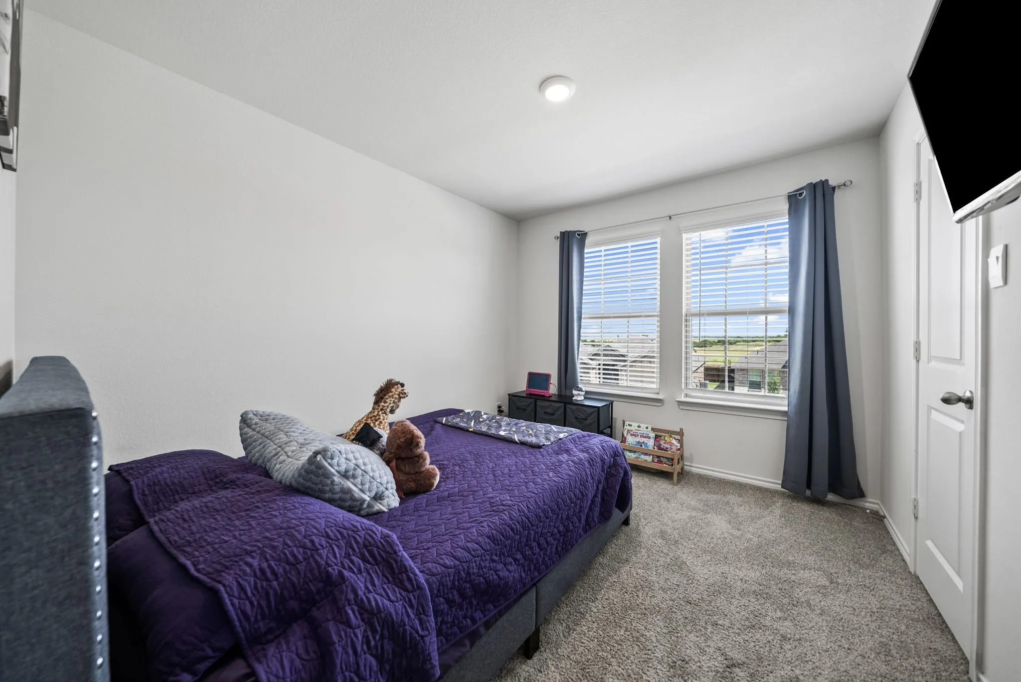 Bright secondary bedroom with dual beds, soft-close carpeting, and large windows with blue thermal curtains providing natural light and privacy