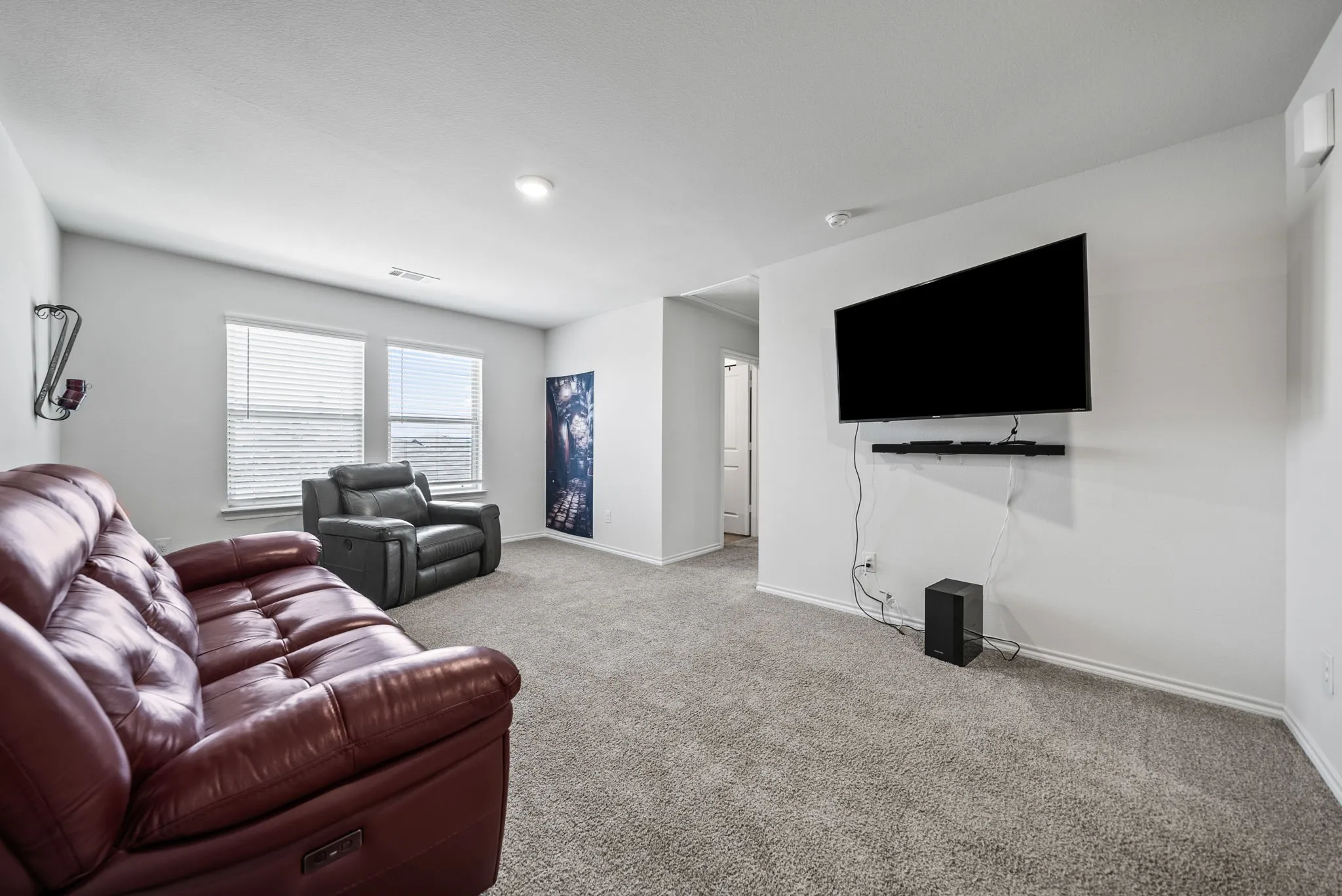 Spacious recreation room featuring wall-mounted flat-screen television with soundbar, burgundy leather reclining sofa, gray upholstered seating, carpeted flooring throughout, recessed lighting, and large windows with blinds providing natural light