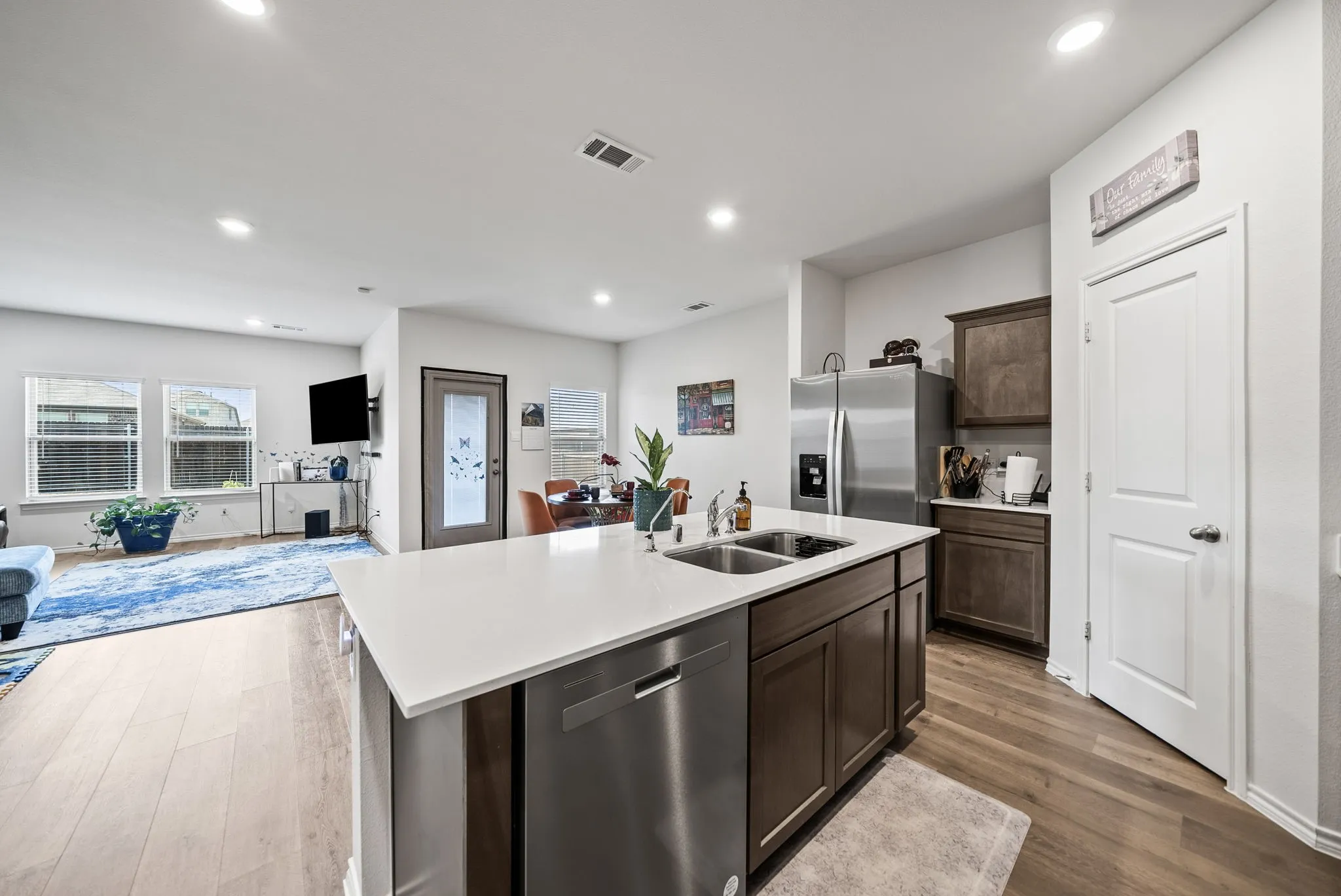 Modern kitchen featuring a large center island with white quartz countertop and dark cabinetry, stainless steel appliances including a refrigerator and range, recessed ceiling lighting, wood-finish flooring, and abundant natural light from multiple windows overlooking the adjacent living space