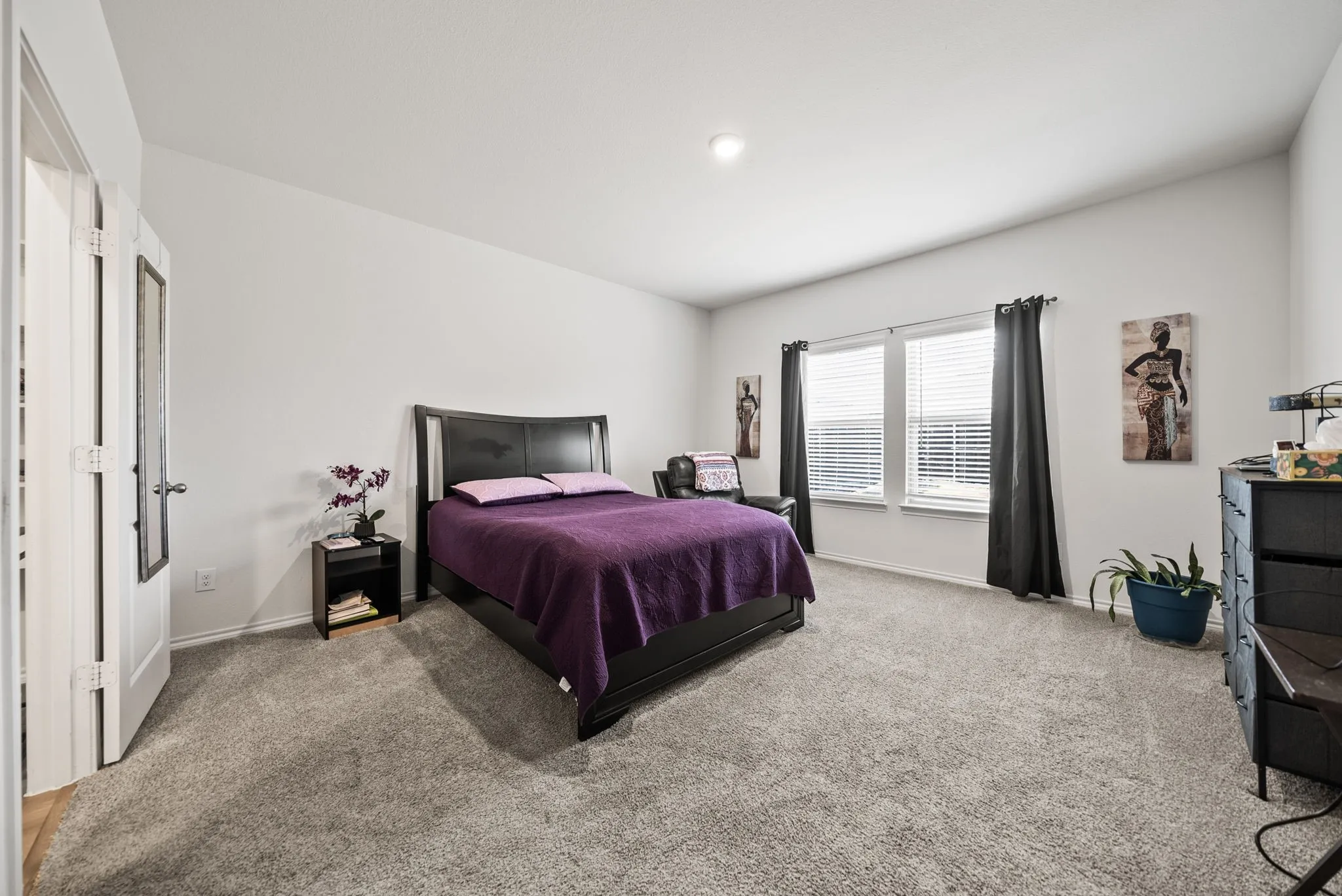 Spacious primary bedroom featuring neutral carpet flooring, large windows with black drapery hardware, a black metal-frame bed, dual closets with white doors, recessed ceiling lighting, and a dark dresser with integrated storage