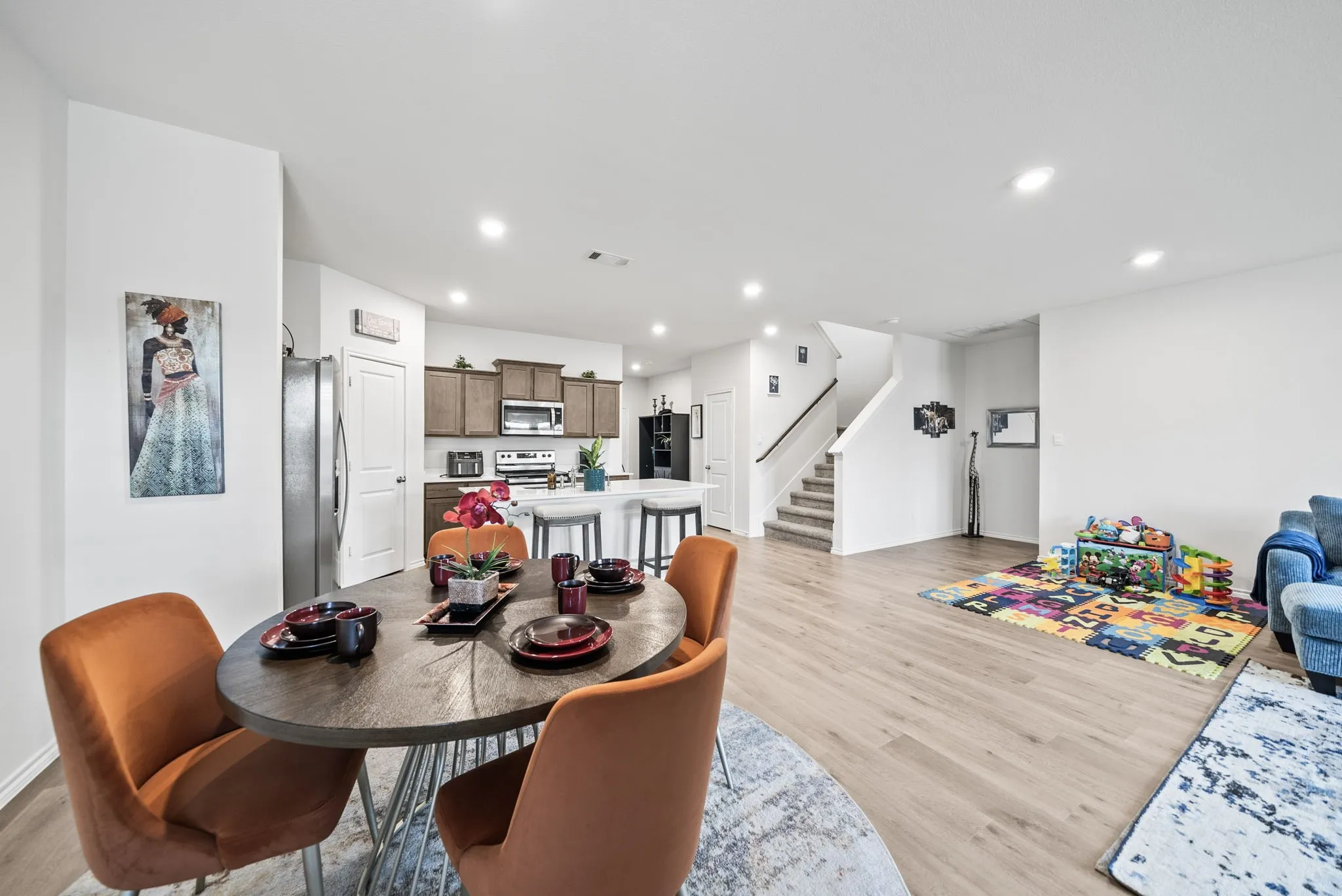 Open-concept living area featuring wood-finish flooring throughout, a round dining table with seating for four, stainless steel refrigerator, white cabinetry with open shelving, recessed lighting, and direct sightlines to a carpeted play space and staircase with white railings