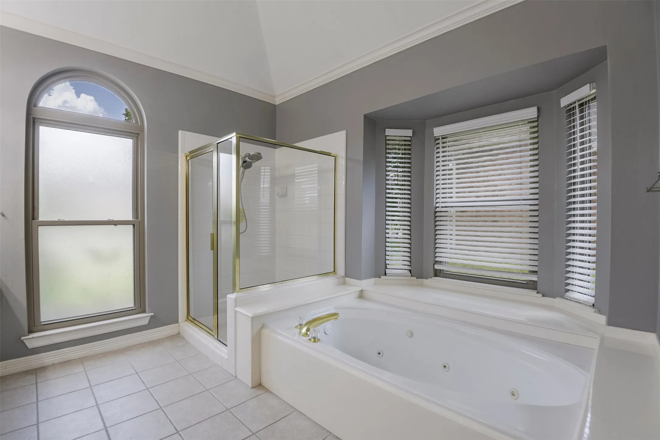 Soaking tub with integrated jets, glass-enclosed shower with brass accents, arched window, bay window, and tile flooring