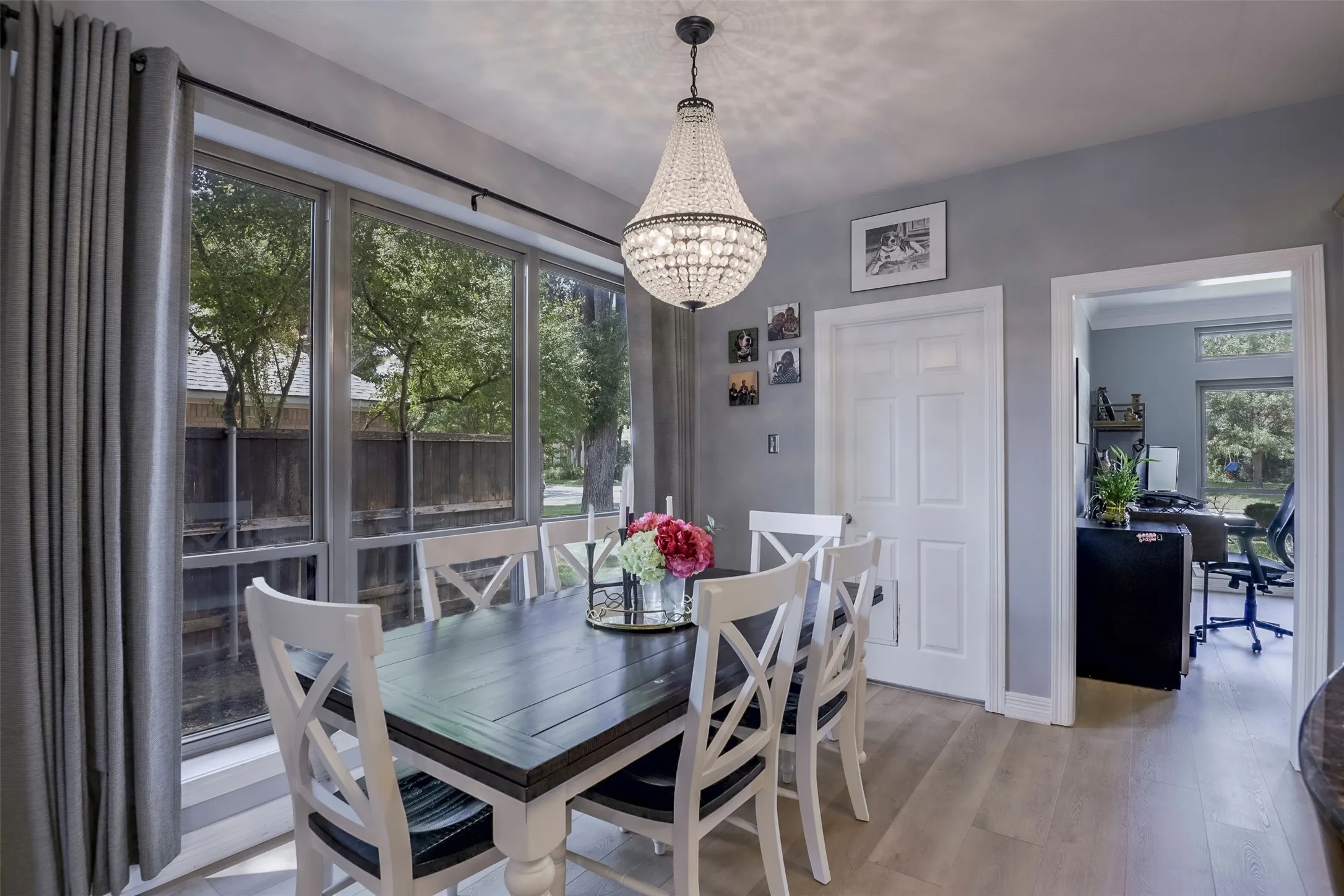 Expansive window wall with grey drapery, central crystal chandelier, light wood-finish flooring, and a white paneled door