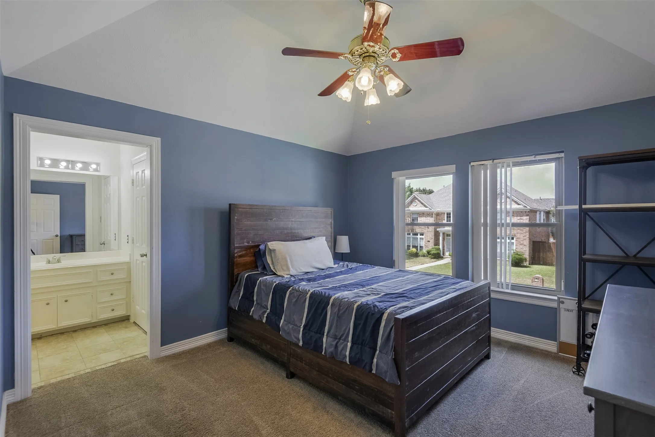 Vaulted ceiling with a ceiling fan, dual windows, and an en-suite bathroom featuring a vanity with a rectangular mirror and sconce lighting