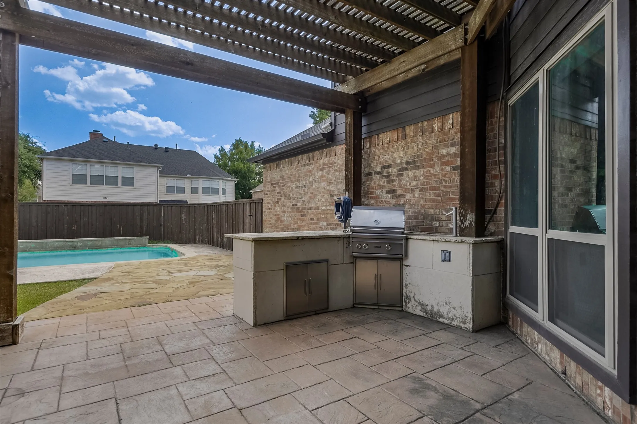 Expansive patio with built-in outdoor kitchen featuring stainless steel appliances and stone countertop, complemented by a pergola overhead and inground swimming pool visible beyond