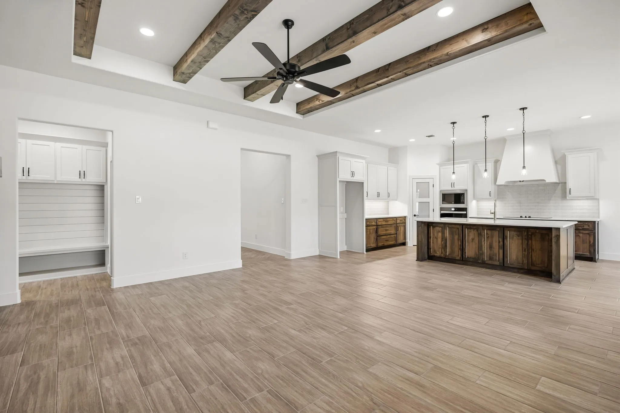 Open-concept living space featuring engineered wood-finish flooring throughout, exposed ceiling beams with bronze ceiling fan, recessed lighting, and white shiplap garage door