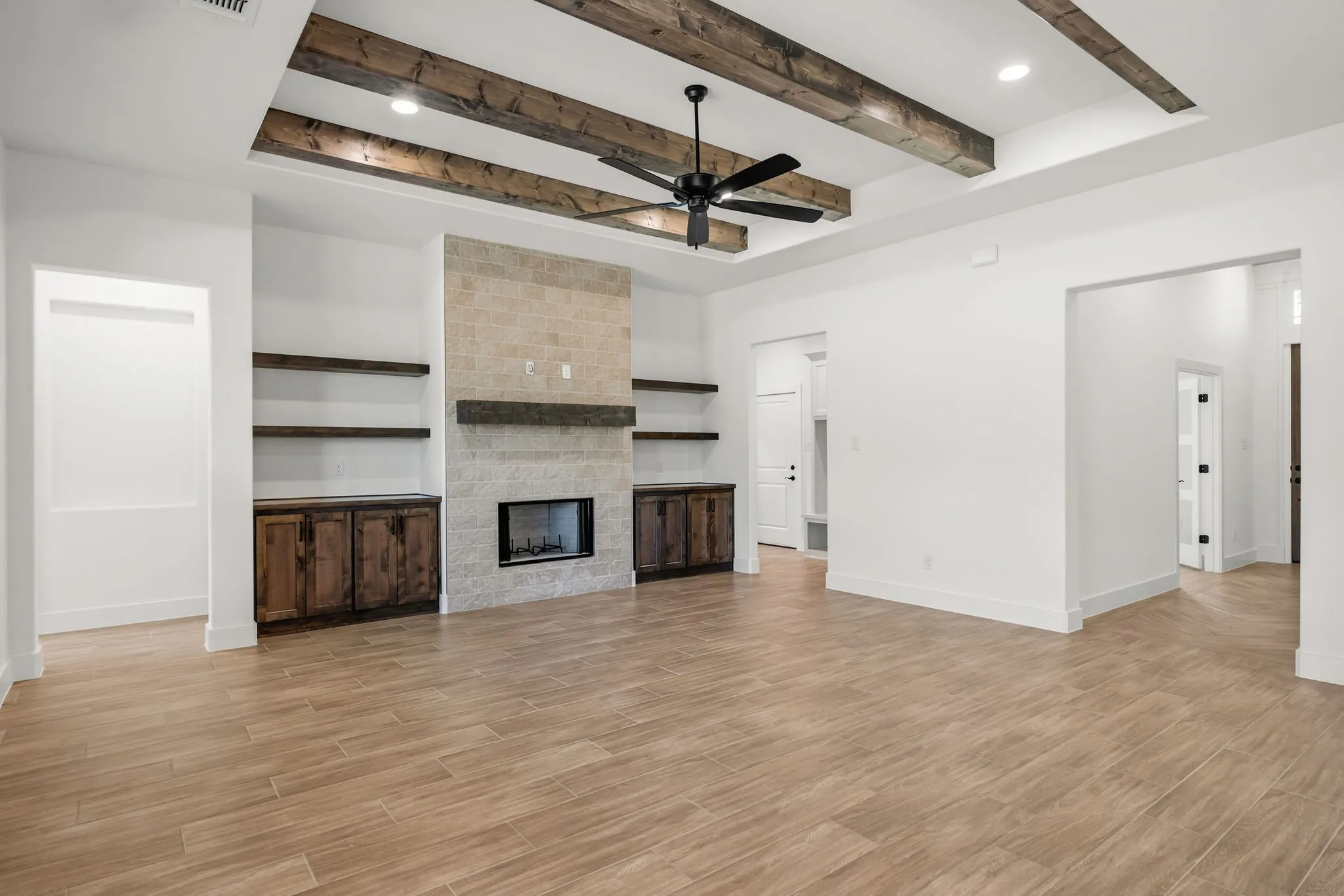 Tray ceiling with exposed wood beams, ceiling fan, fireplace with stone surround, wood-finish flooring, and built-in shelving with lower cabinetry